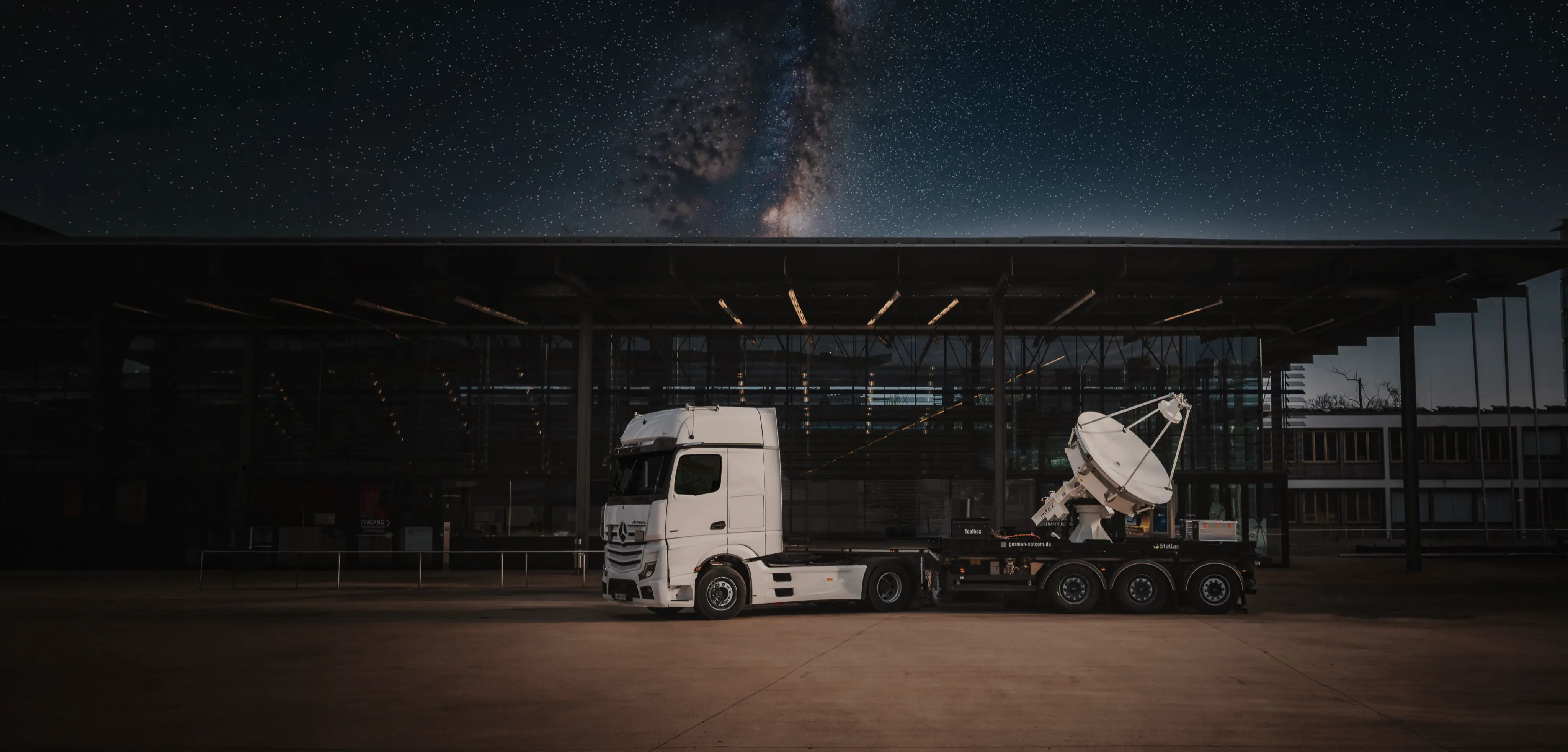 White semi-truck parked in front of a modern glass building at night, with a large satellite dish mounted on its trailer and a starry night sky visible above.
