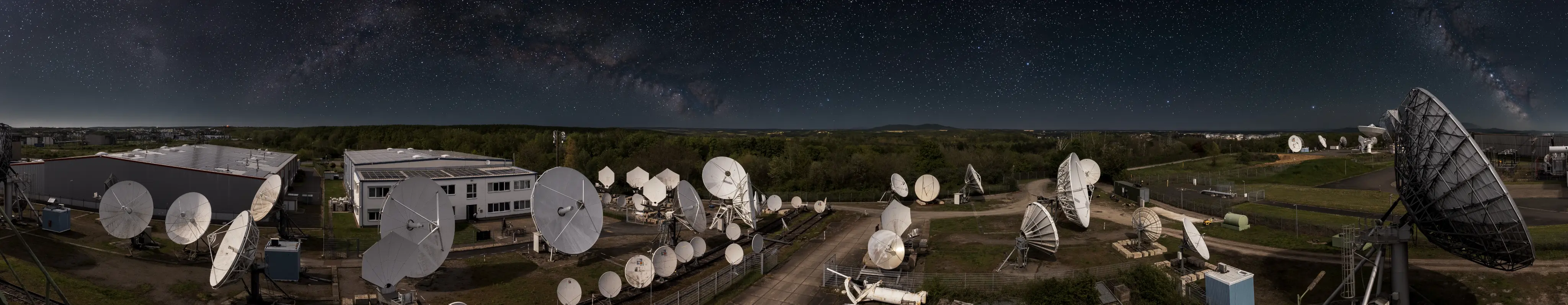 Panoramic night view of a satellite ground station with numerous large white satellite dishes arranged in rows near buildings, under a starry sky with the Milky Way visible across the horizon.