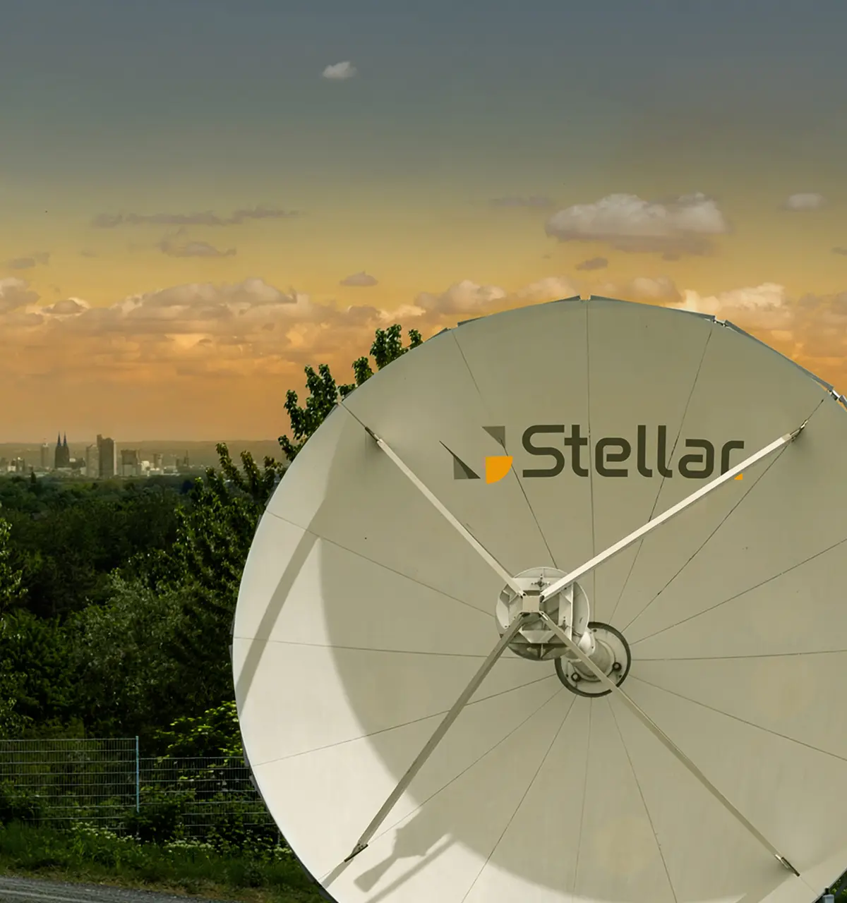 Large white satellite dish with the word 'Stellar' on it, set against a backdrop of green trees and a city skyline under a cloudy orange-yellow sky at sunset.