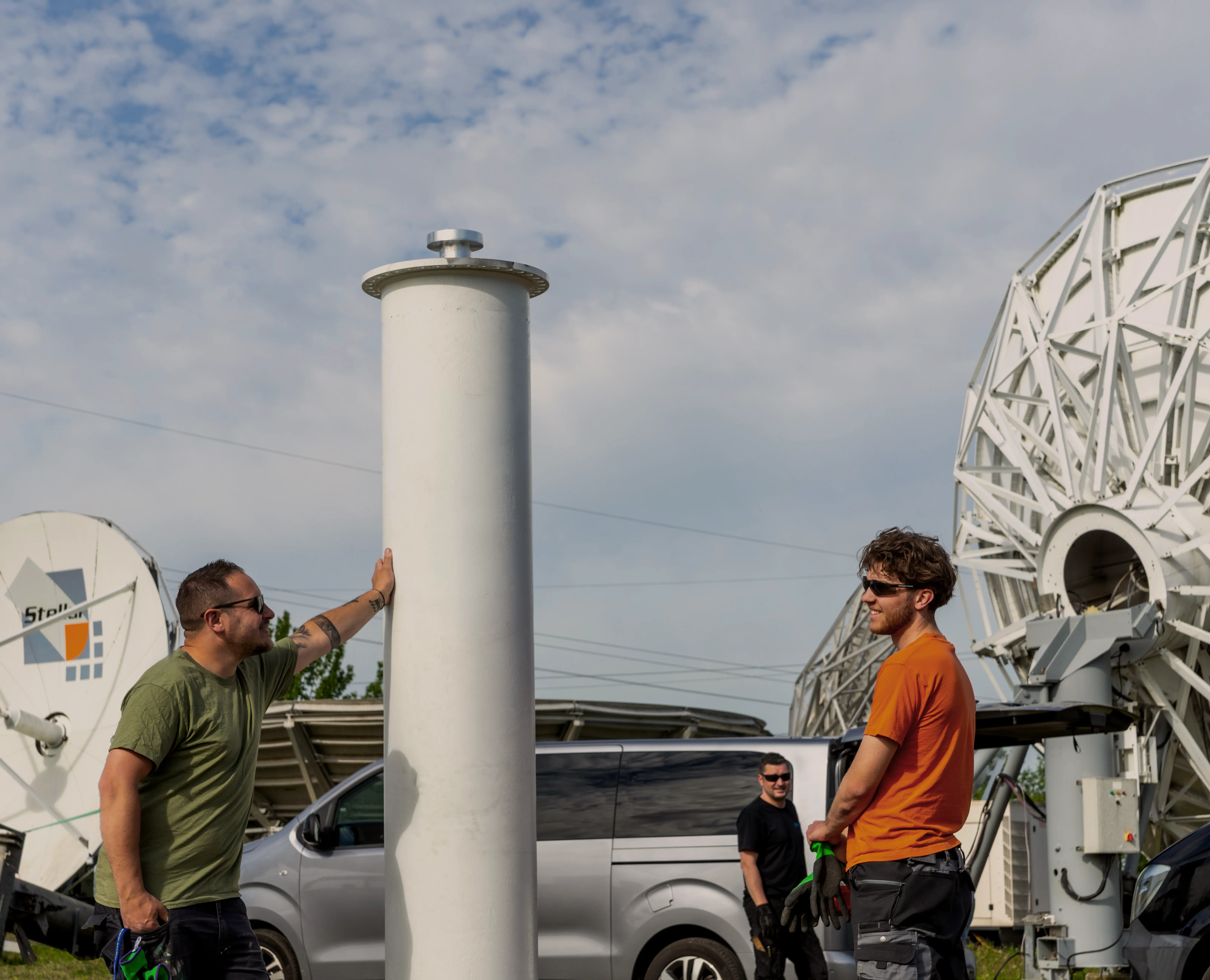 Three men wearing sunglasses and work gloves stand near large satellite dishes and a silver van, with one man leaning on a tall white cylindrical object under a partly cloudy sky.