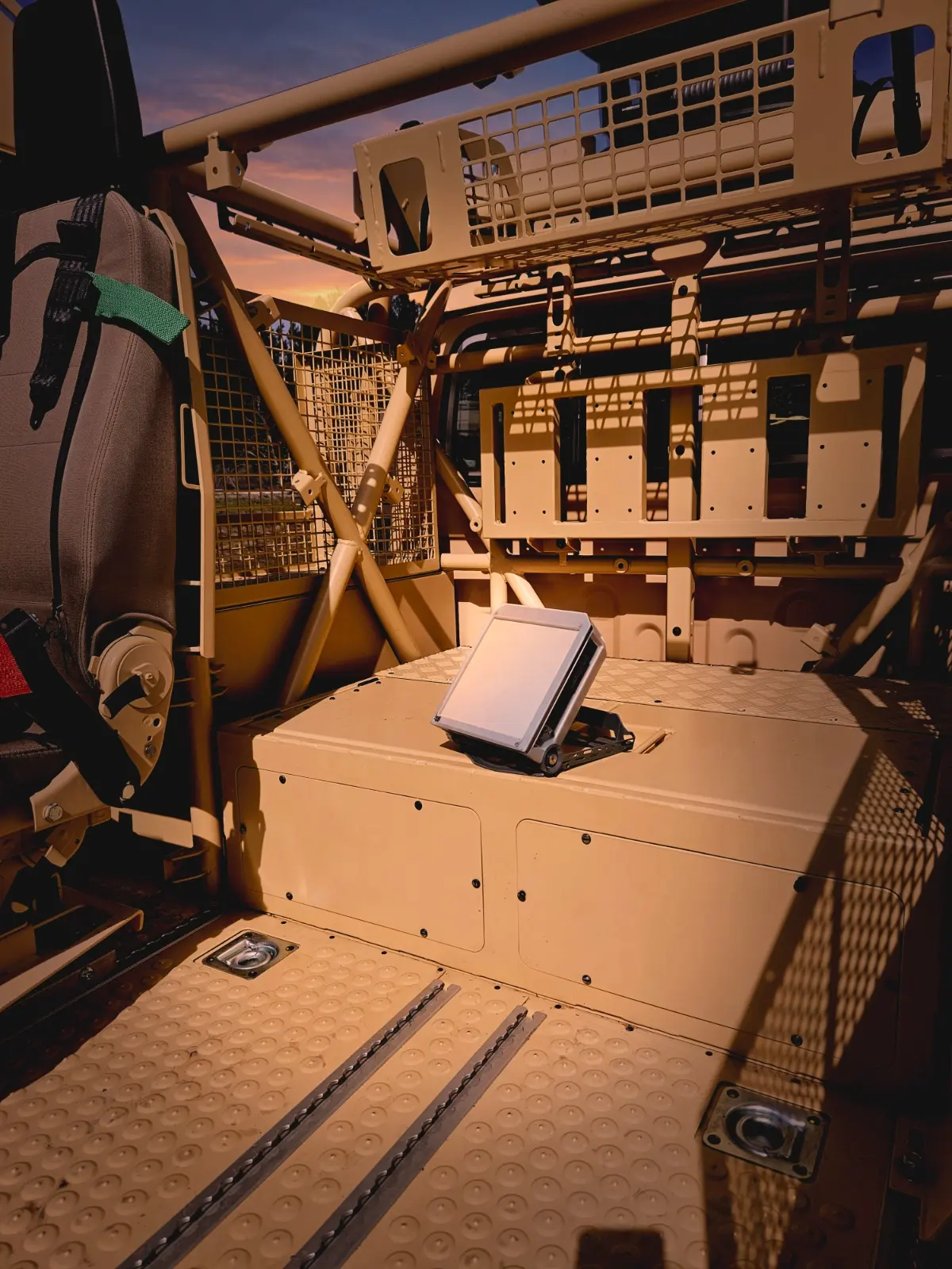 Interior of a tan-colored utility or military vehicle with metal mesh panels, structural bars, a seat with a green strap, and a small elevated light or device mounted on a flat surface inside the vehicle.