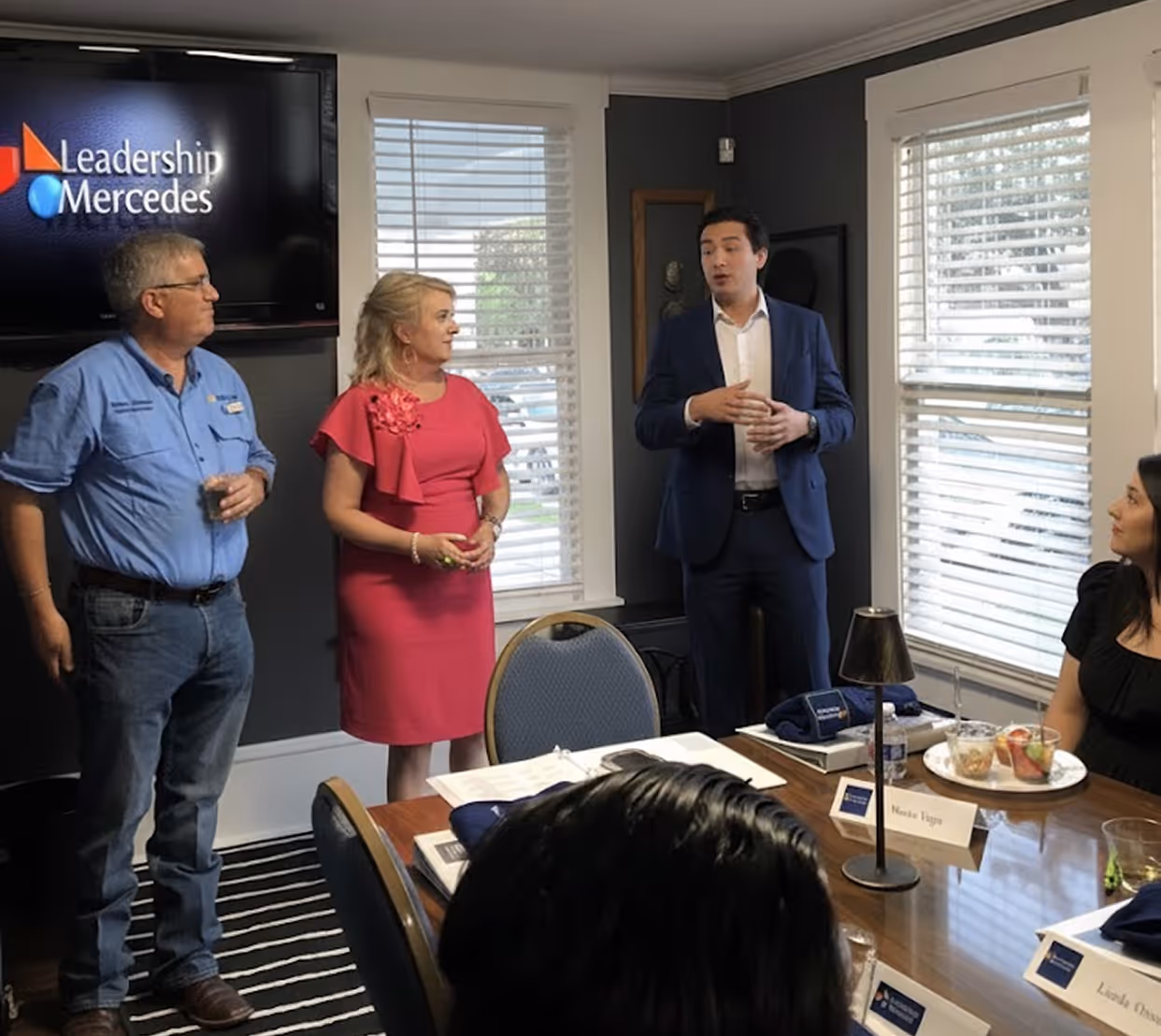 Three people standing and talking in a meeting room with Leadership Mercedes logo on a screen, others seated at a conference table.