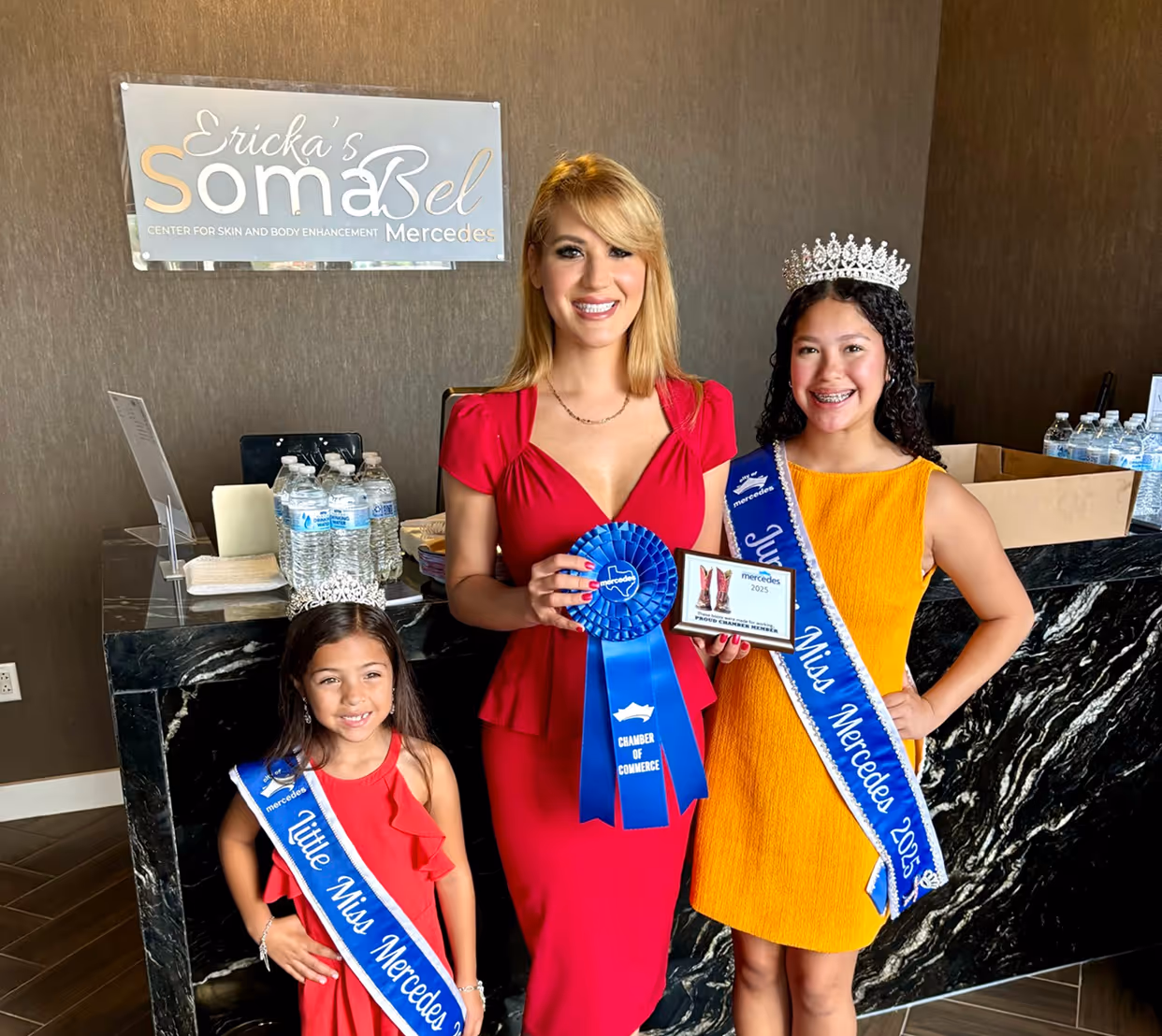 Three smiling females wearing pageant sashes and crowns, one holding a blue Chamber of Commerce ribbon and plaque inside a room with a black marble counter.