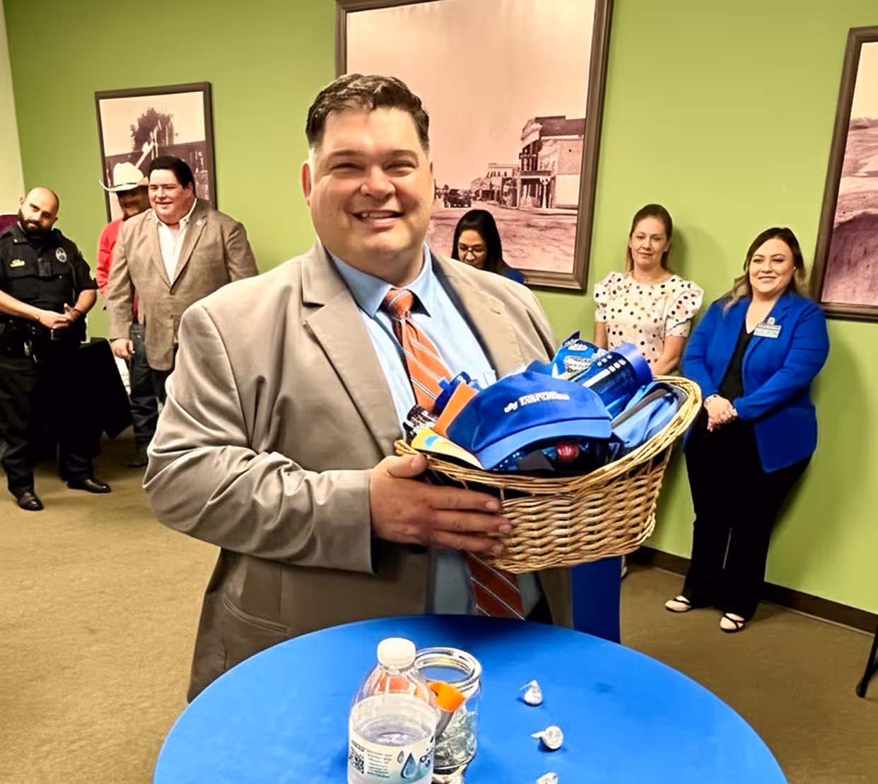 Smiling man in tan suit holds a basket with blue promotional items, with people standing and smiling in the background.