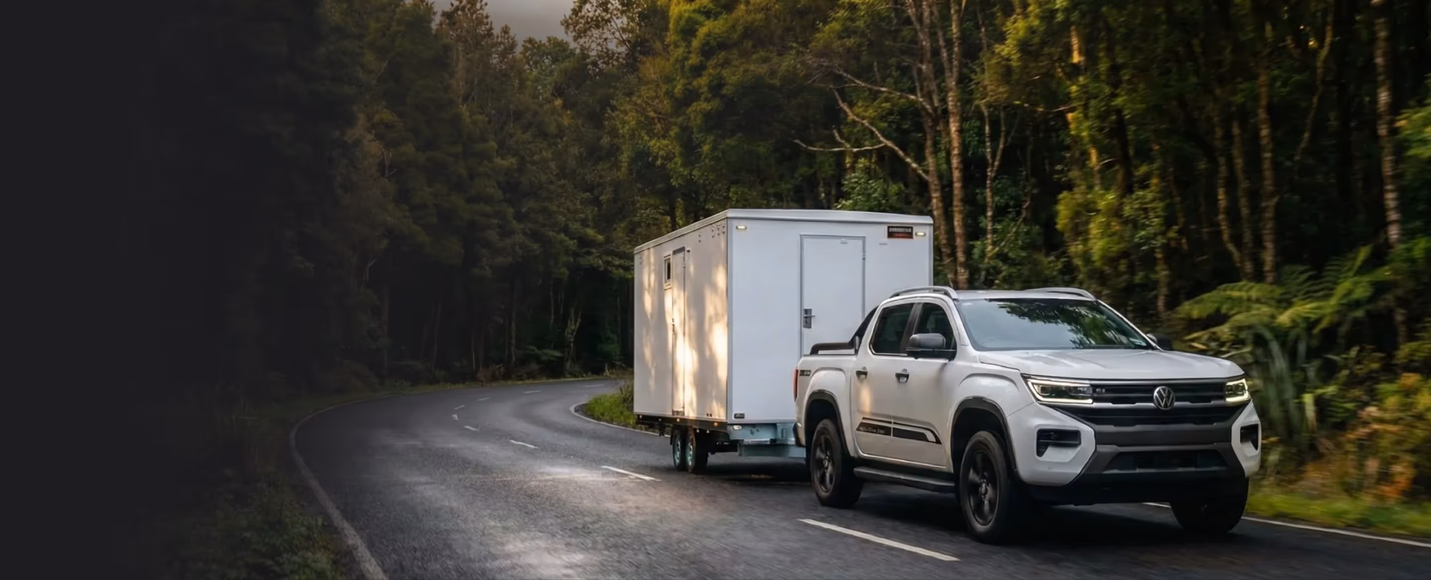 White pickup truck towing a white portable restroom trailer on a curved forest road