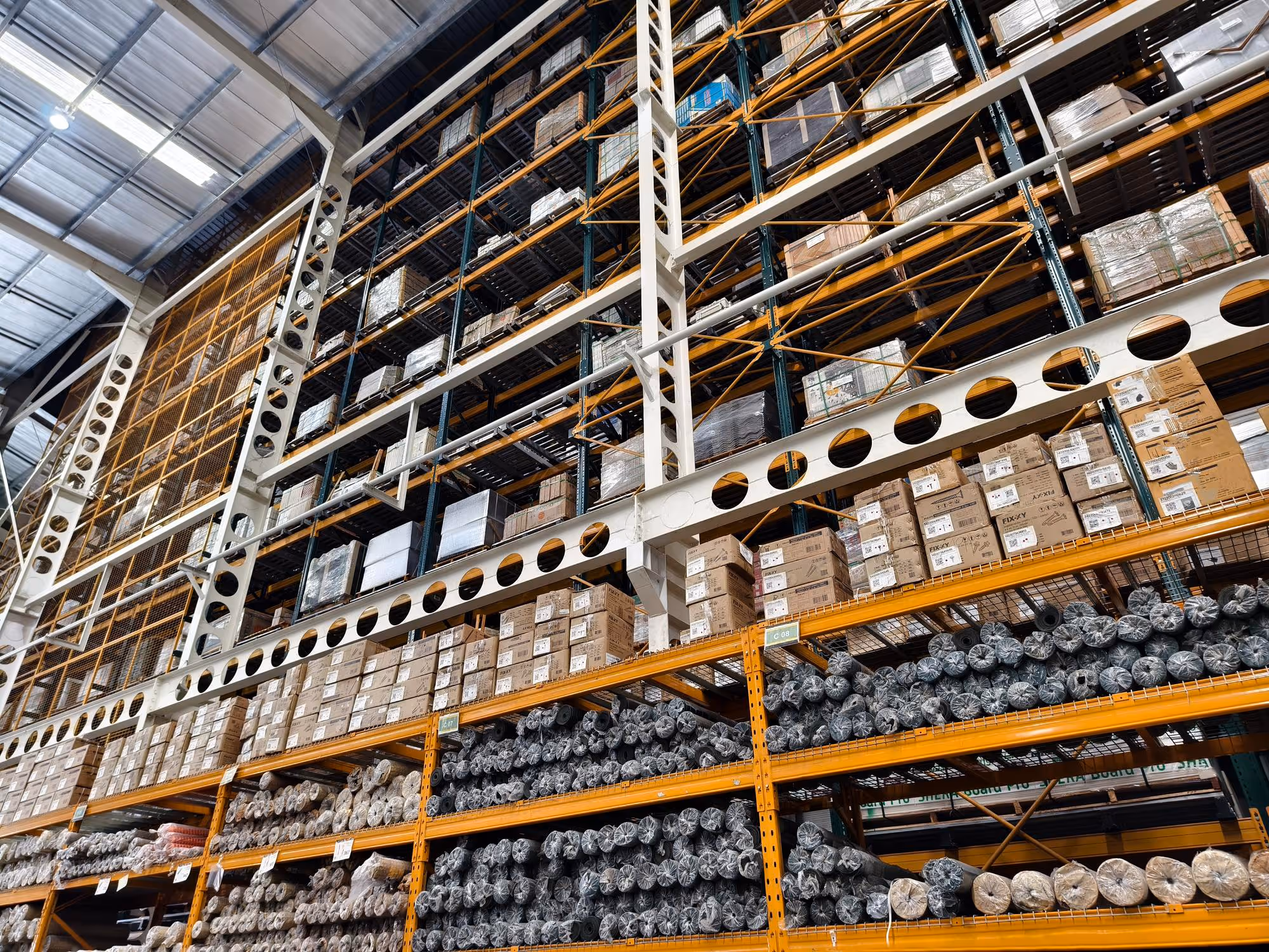 Warehouse shelves filled with stacked cardboard boxes and cylindrical items wrapped in plastic