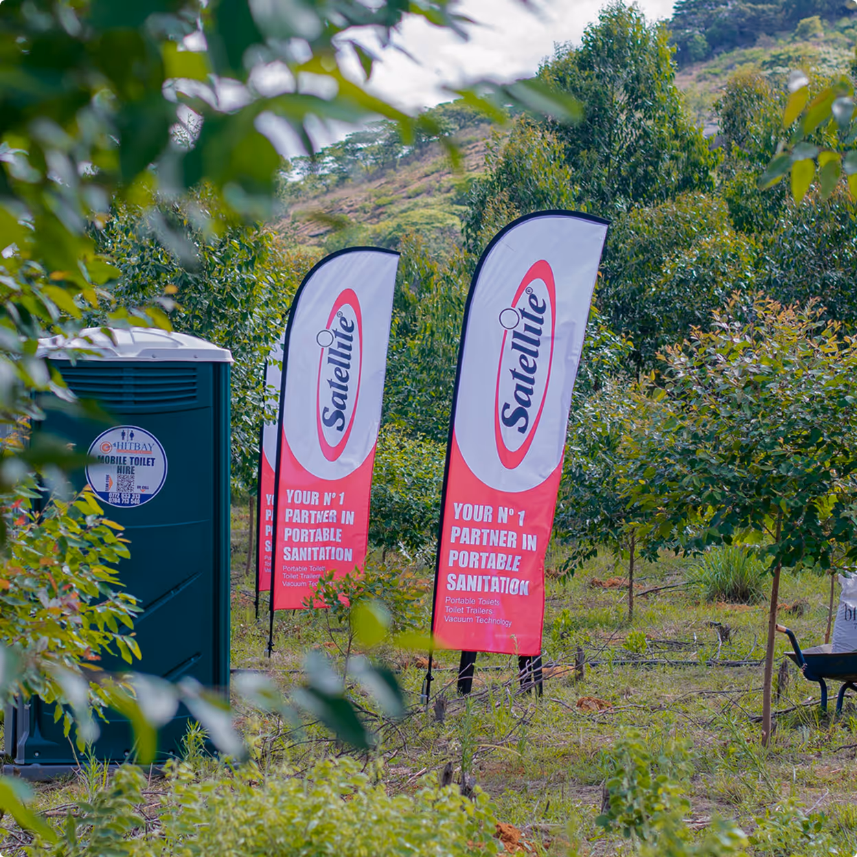 Green portable restroom next to three Satellite Industries branded flags in a green outdoor setting