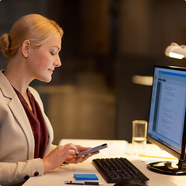 Woman in beige blazer using smartphone at desk with computer monitor and keyboard