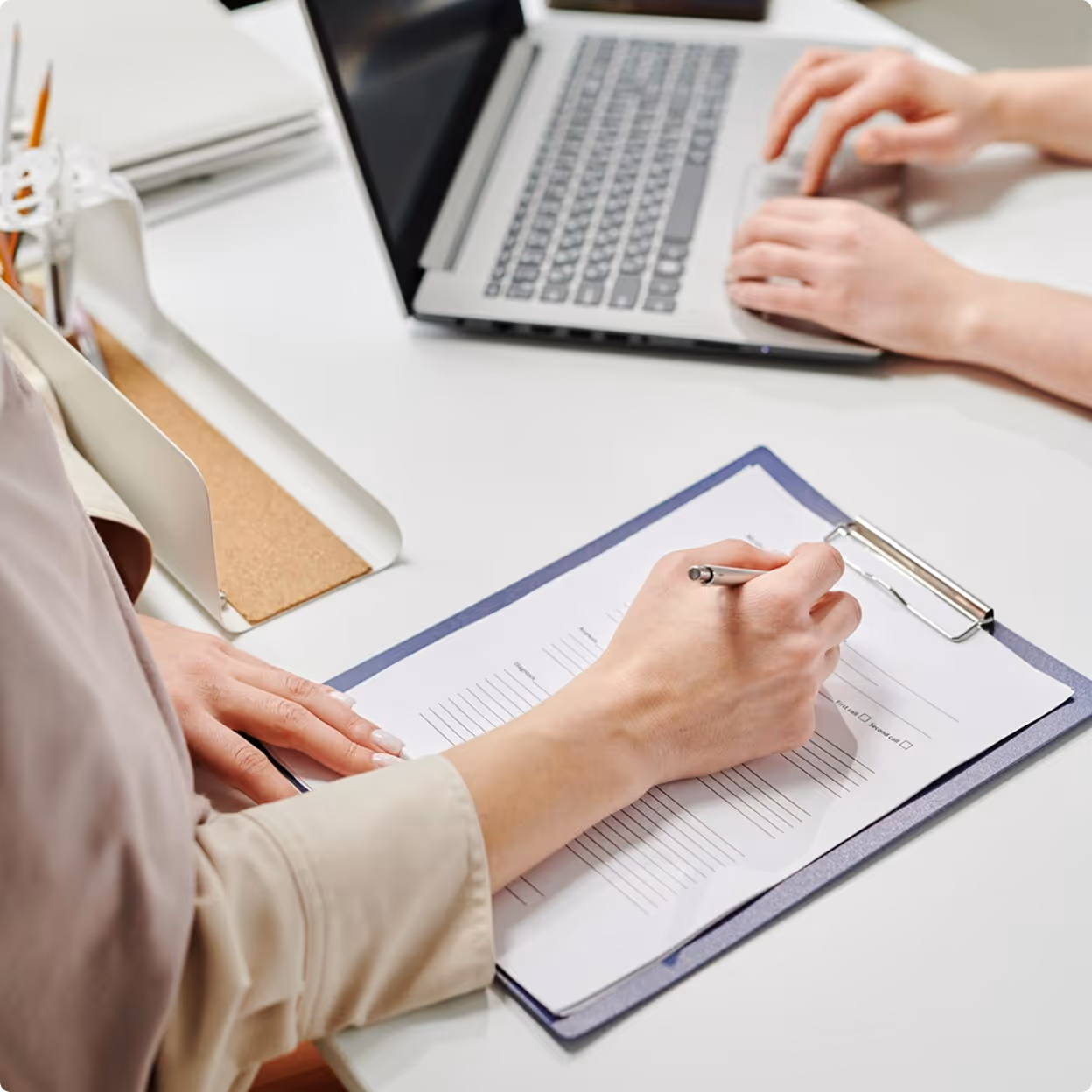 Person filling out form on clipboard next to laptop keyboard on white desk