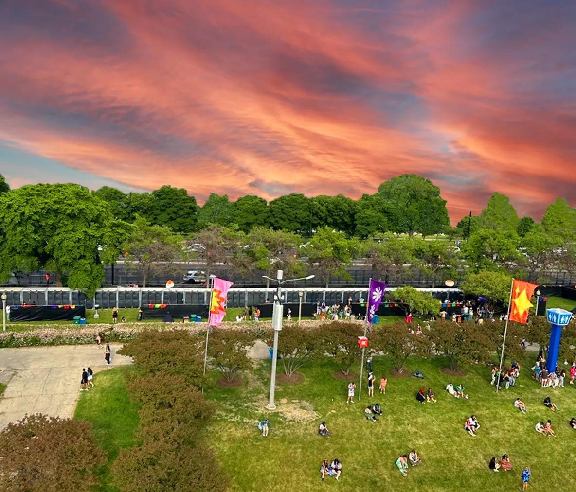 Outdoor event with groups of people sitting on grass near colorful flags and tall trees under a vibrant sunset sky