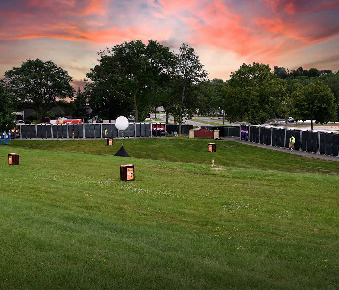 Row of black portable restrooms set along road at outdoor event with tree-lined background and grassy foreground at sunset