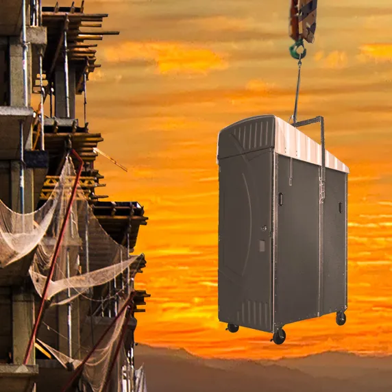 Dark gray portable restroom with white roof being lifted by crane at construction site during sunset