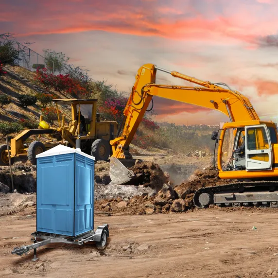 Blue portable restroom trailer on construction site with excavator and grader at sunset