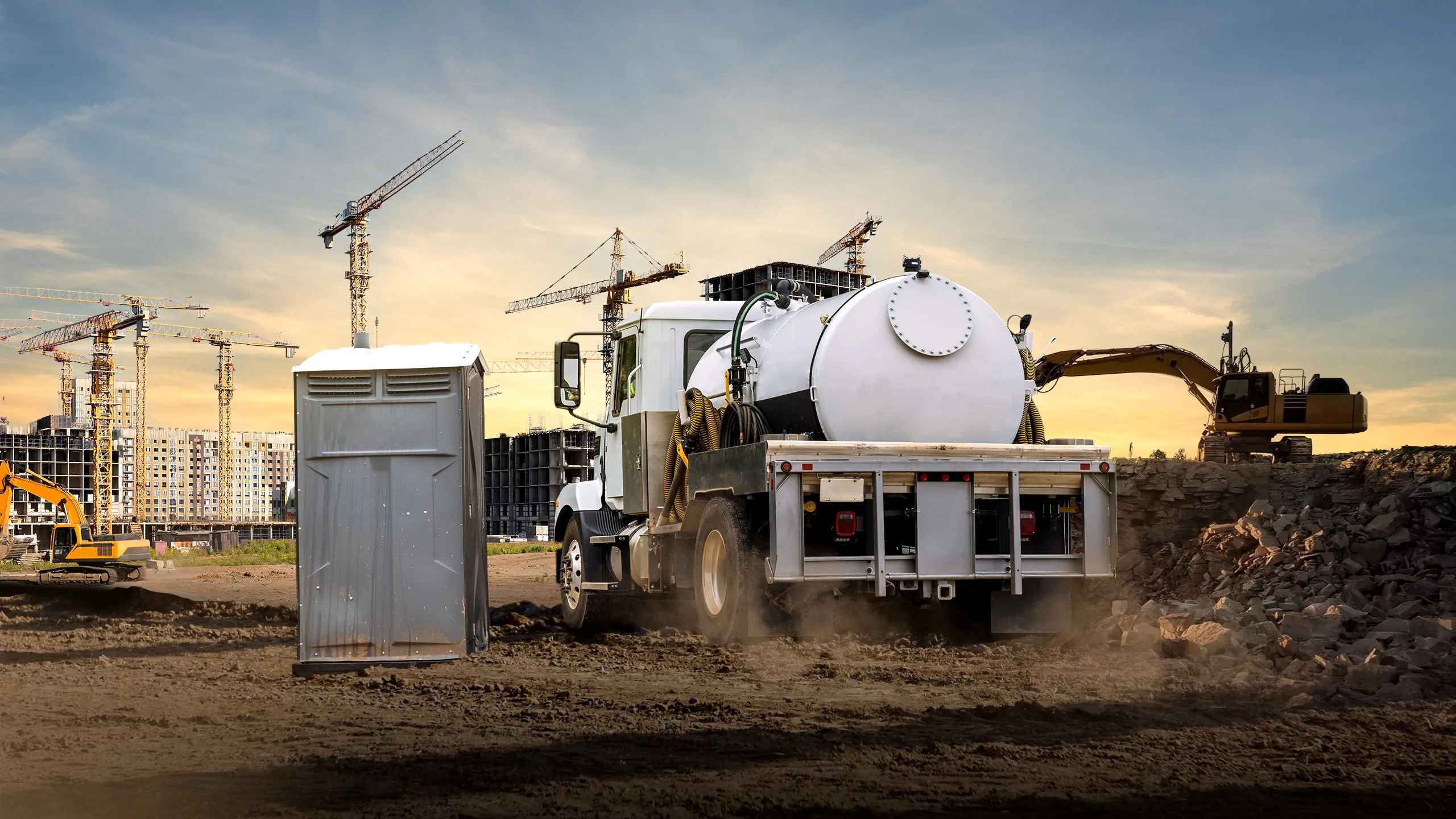 Vacuum service truck rear view next to portable restroom on construction site with cranes and excavators in background