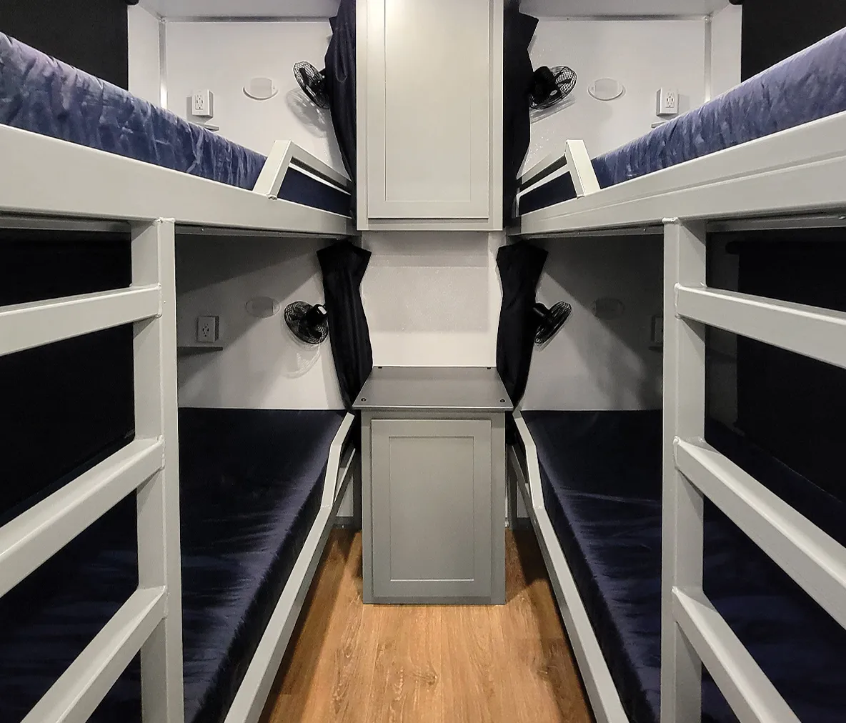 Four gray bunk beds with blue mattresses and individual fans in compact room with wood floor