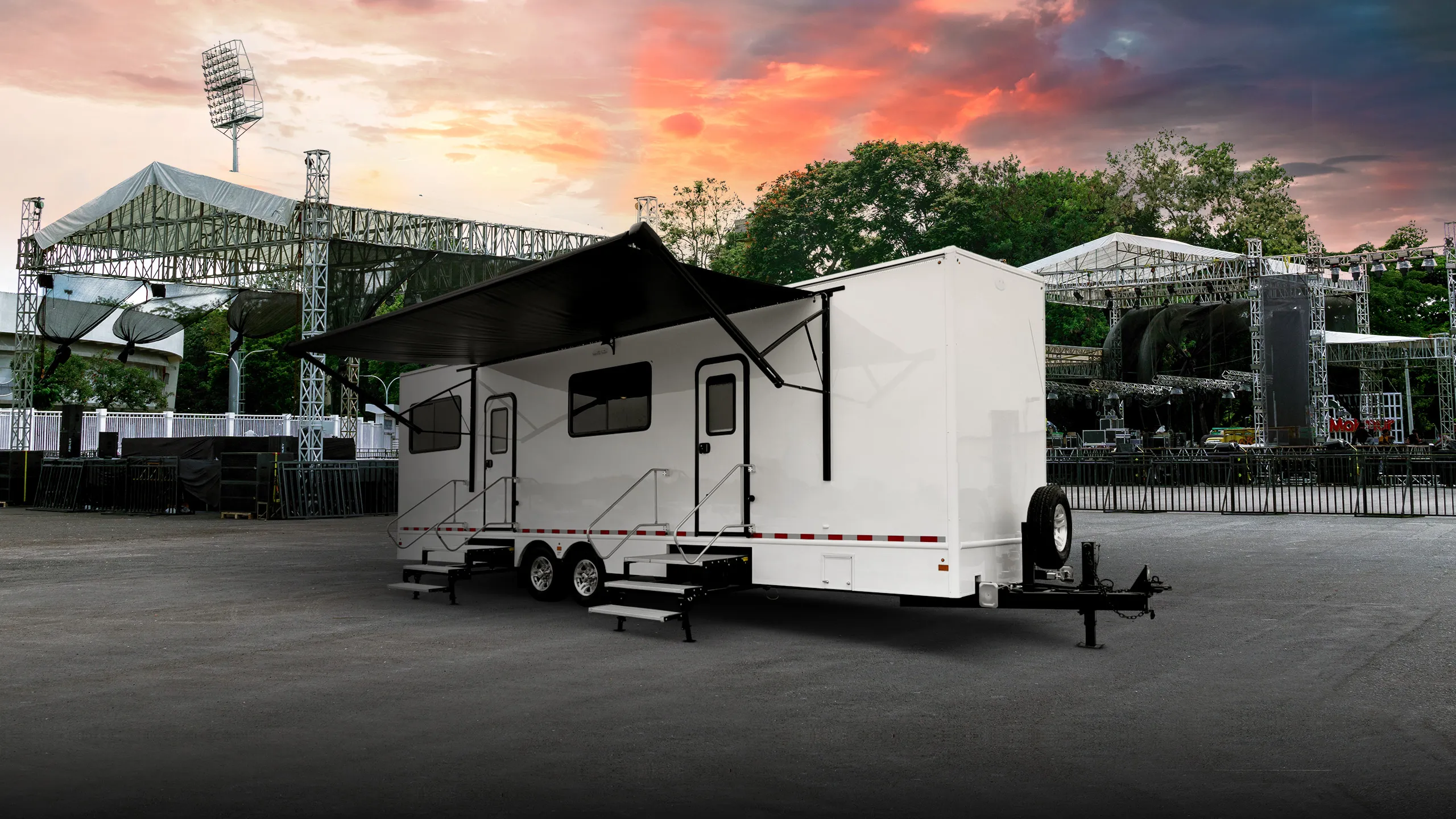 White restroom trailer with extended black awning parked near outdoor event stage at sunset