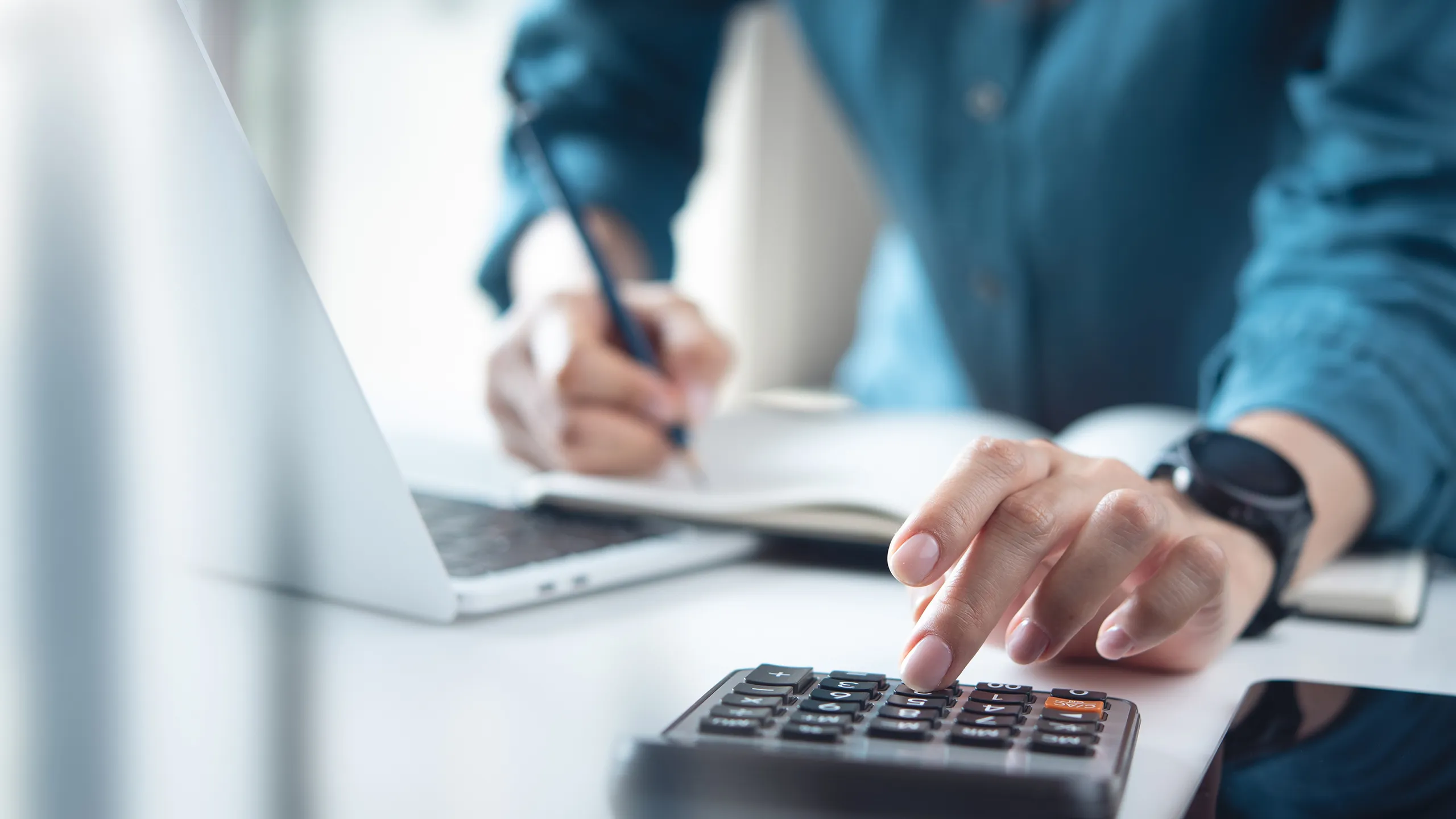 Person pressing calculator button while taking notes near laptop on desk