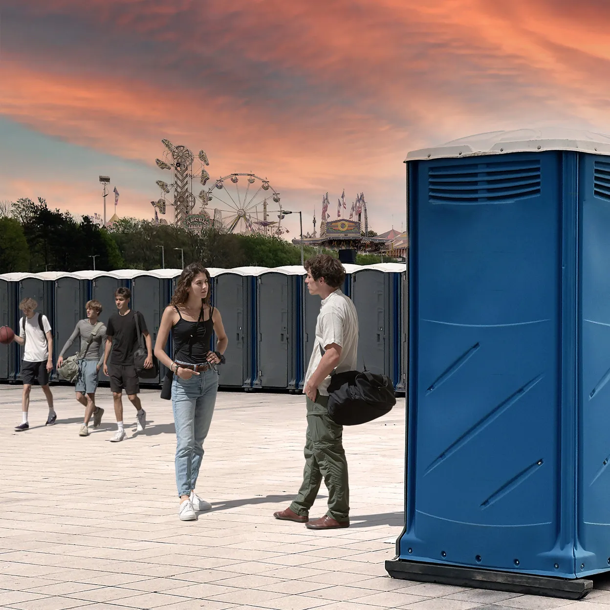 Blue portable restrooms lined up outdoors at amusement park with people walking and talking nearby during sunset