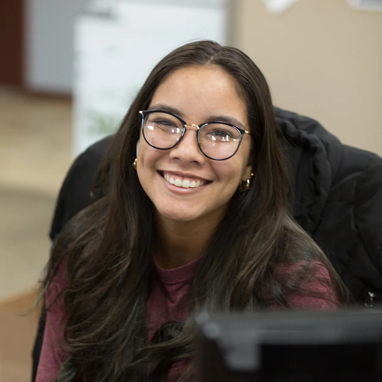 Young woman with long dark hair and glasses smiling indoors