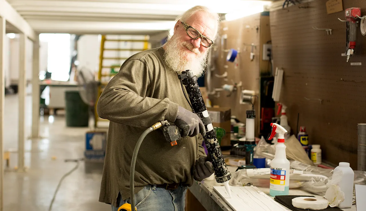 Man with white beard and safety glasses using caulking gun in workshop
