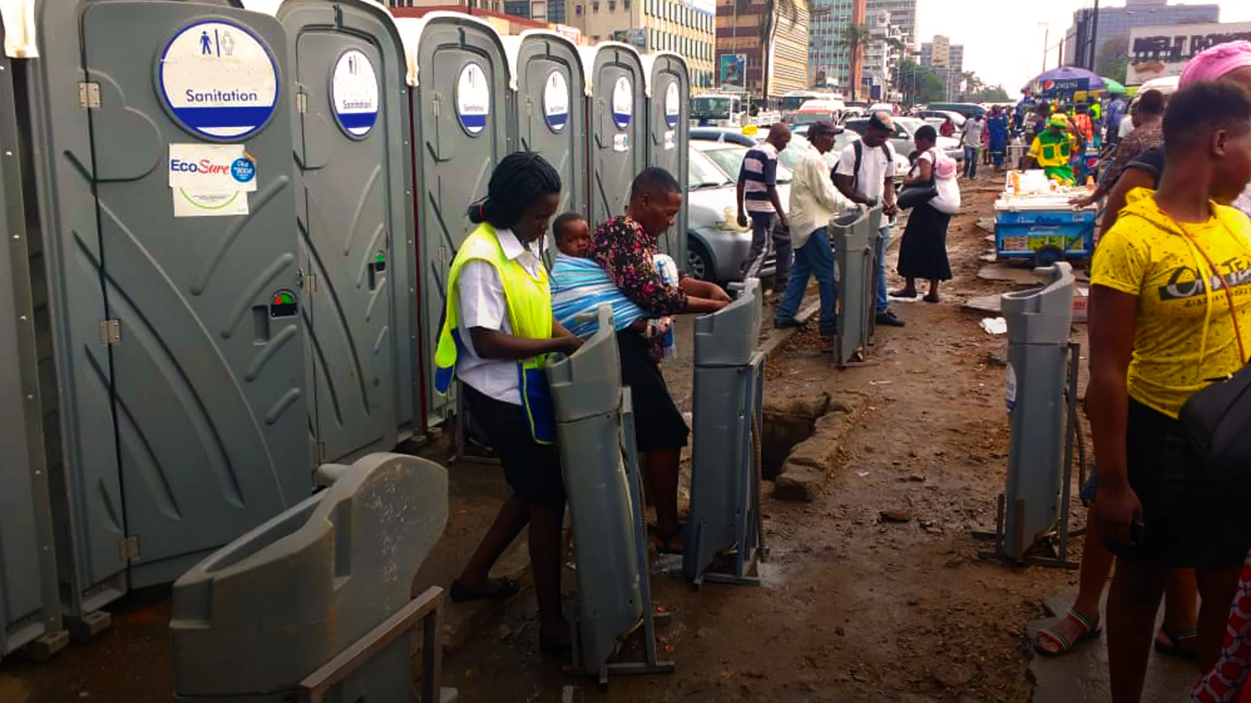 Row of gray portable restrooms with multiple people using portable handwash stations on busy urban street