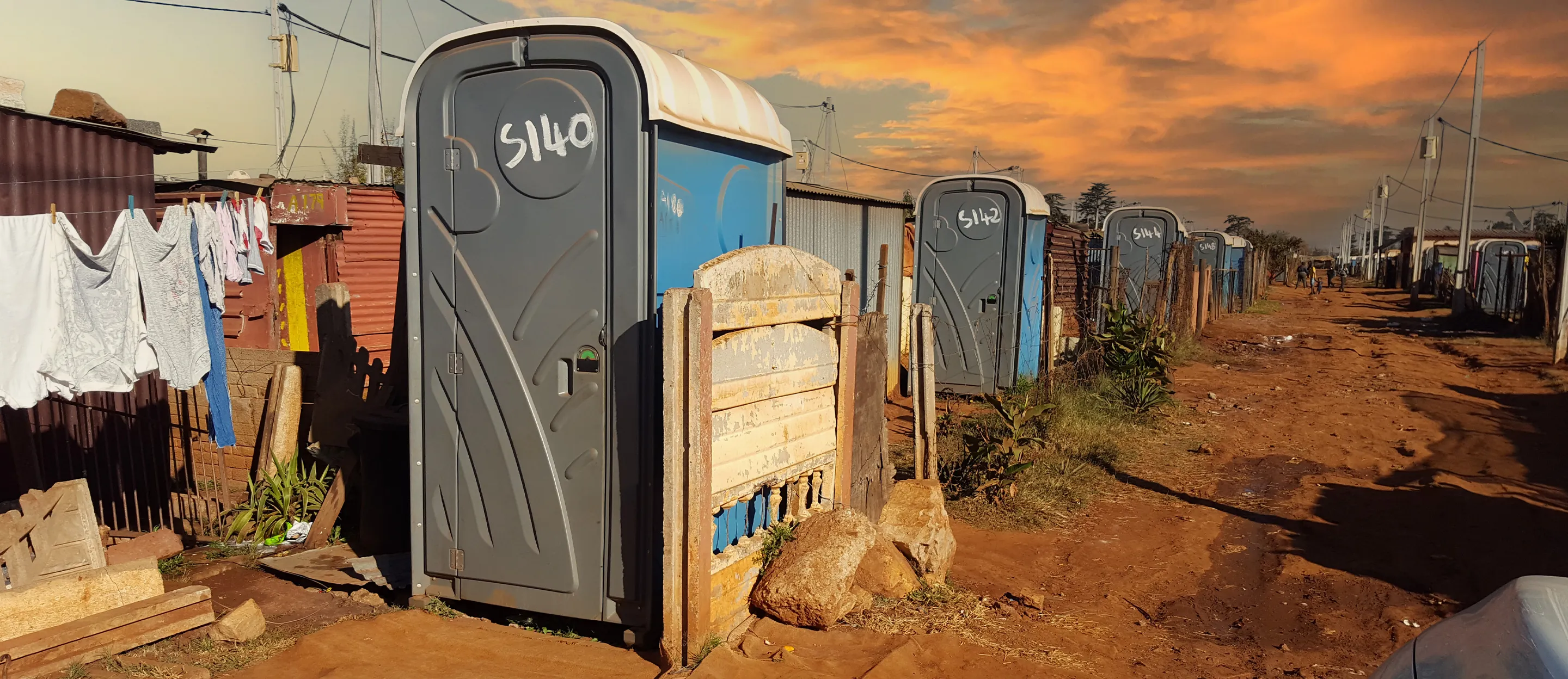 Row of blue portable restrooms with gray doors and white numbers on a dirt road at sunset