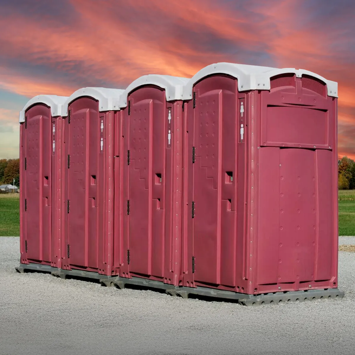 Five maroon portable restrooms with white roofs lined up on gravel at sunset