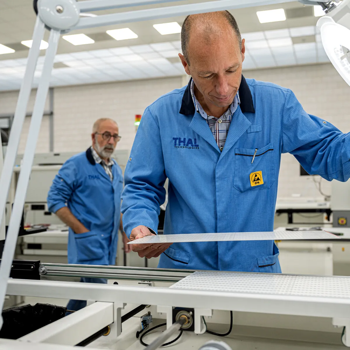 Two factory workers in blue EPA-approved lab coats inspecting a flat perforated panel in a manufacturing setting