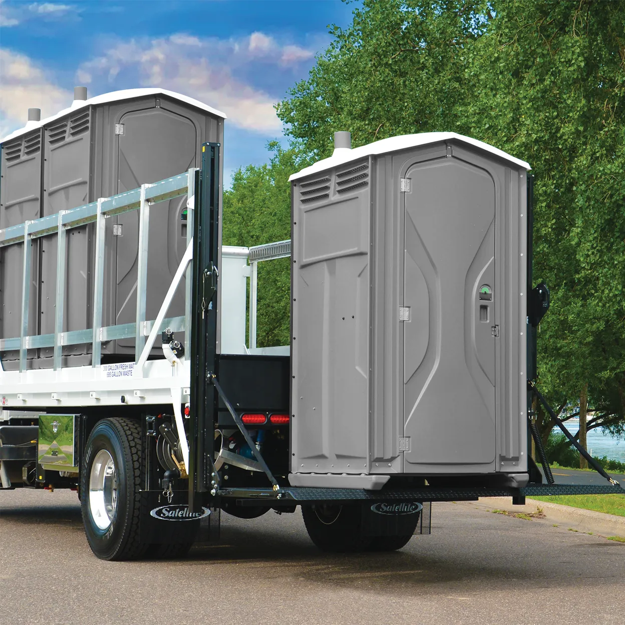 Gray portable restrooms loaded on back of flatbed truck parked on street with trees in background