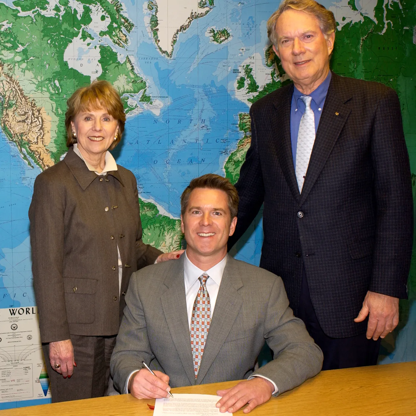 Man in gray suit signing document at table with woman and man standing behind him in front of large world map