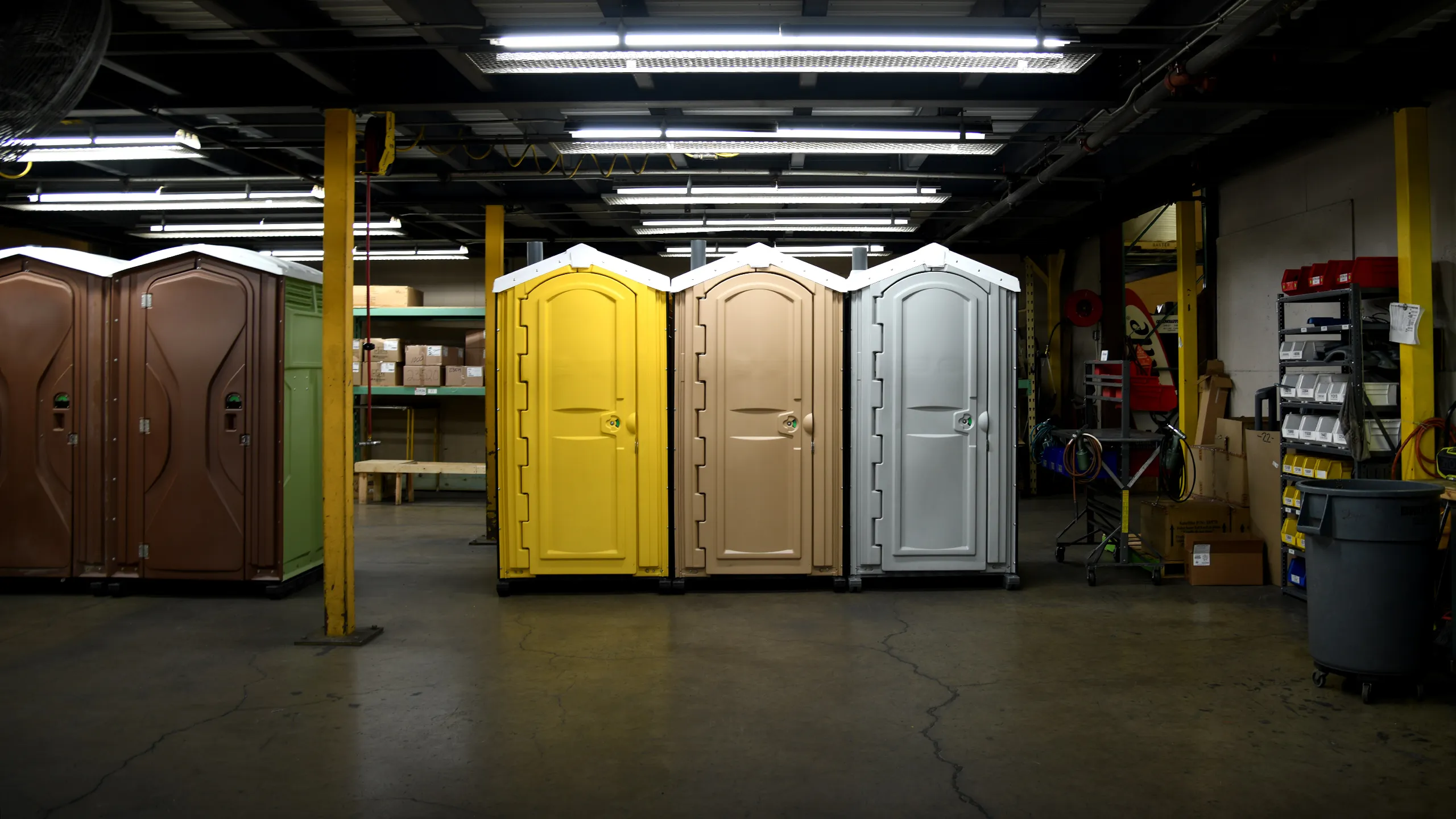 Three portable restrooms in yellow, beige, and gray inside a warehouse with industrial shelving and lighting