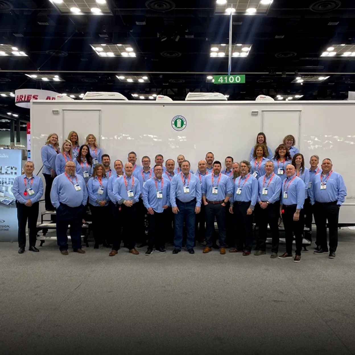 Group of people wearing light blue shirts and conference badges standing in front of a large white trailer at an indoor event