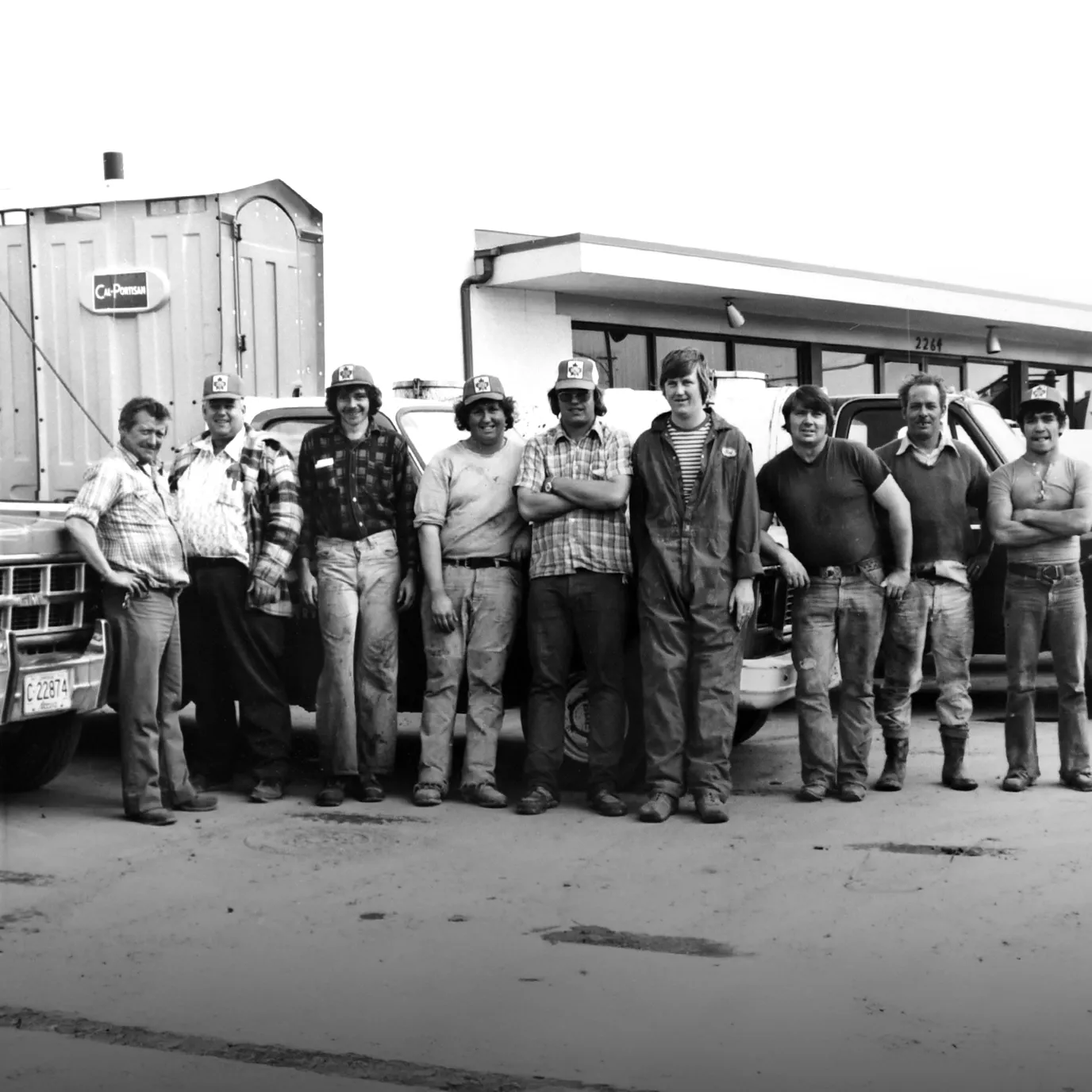 Group of nine men in work clothes and hats standing in front of work trucks outside a building in 1988