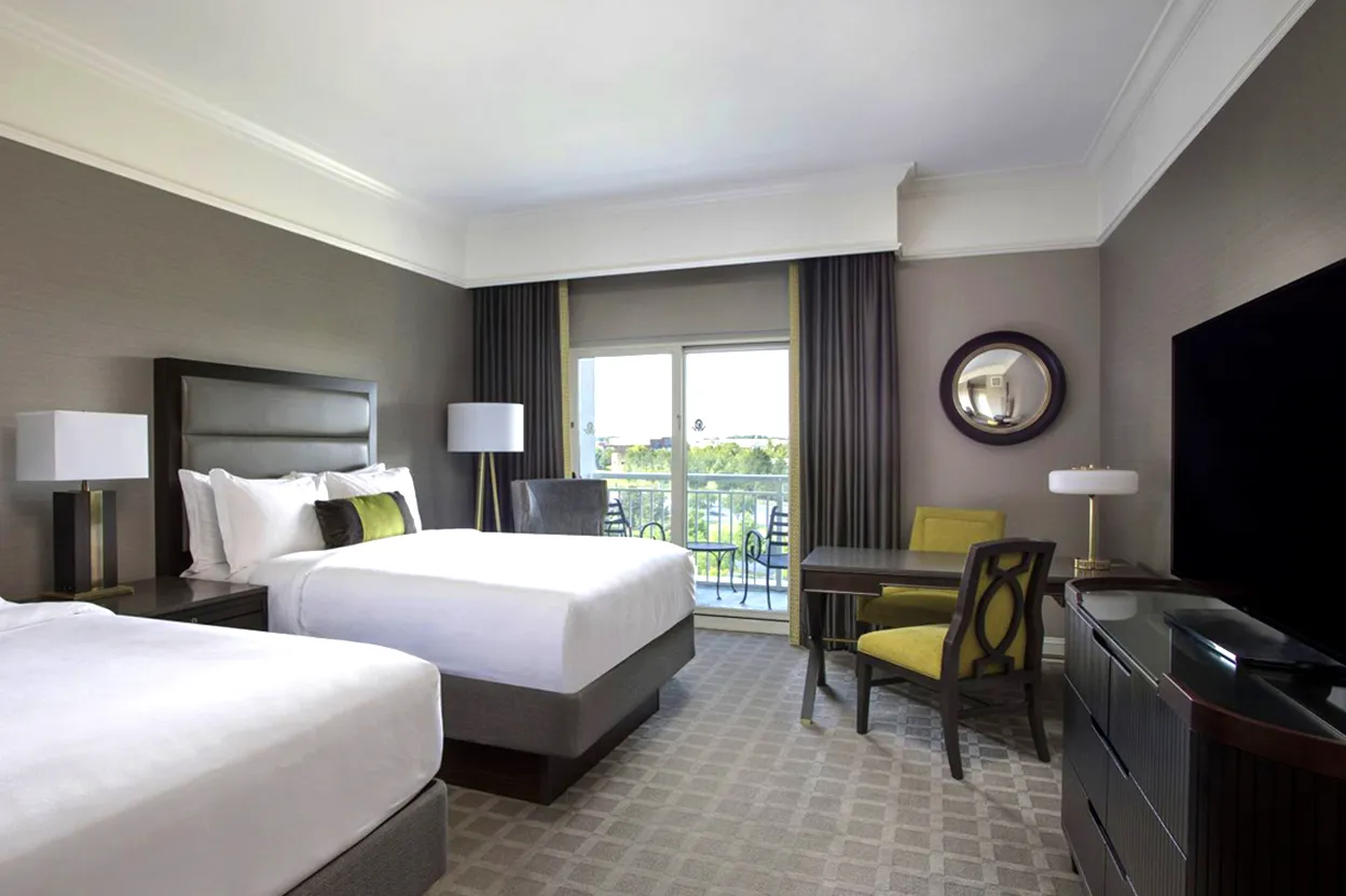 Hotel room with two beds, table with yellow chairs, and balcony door with view outside