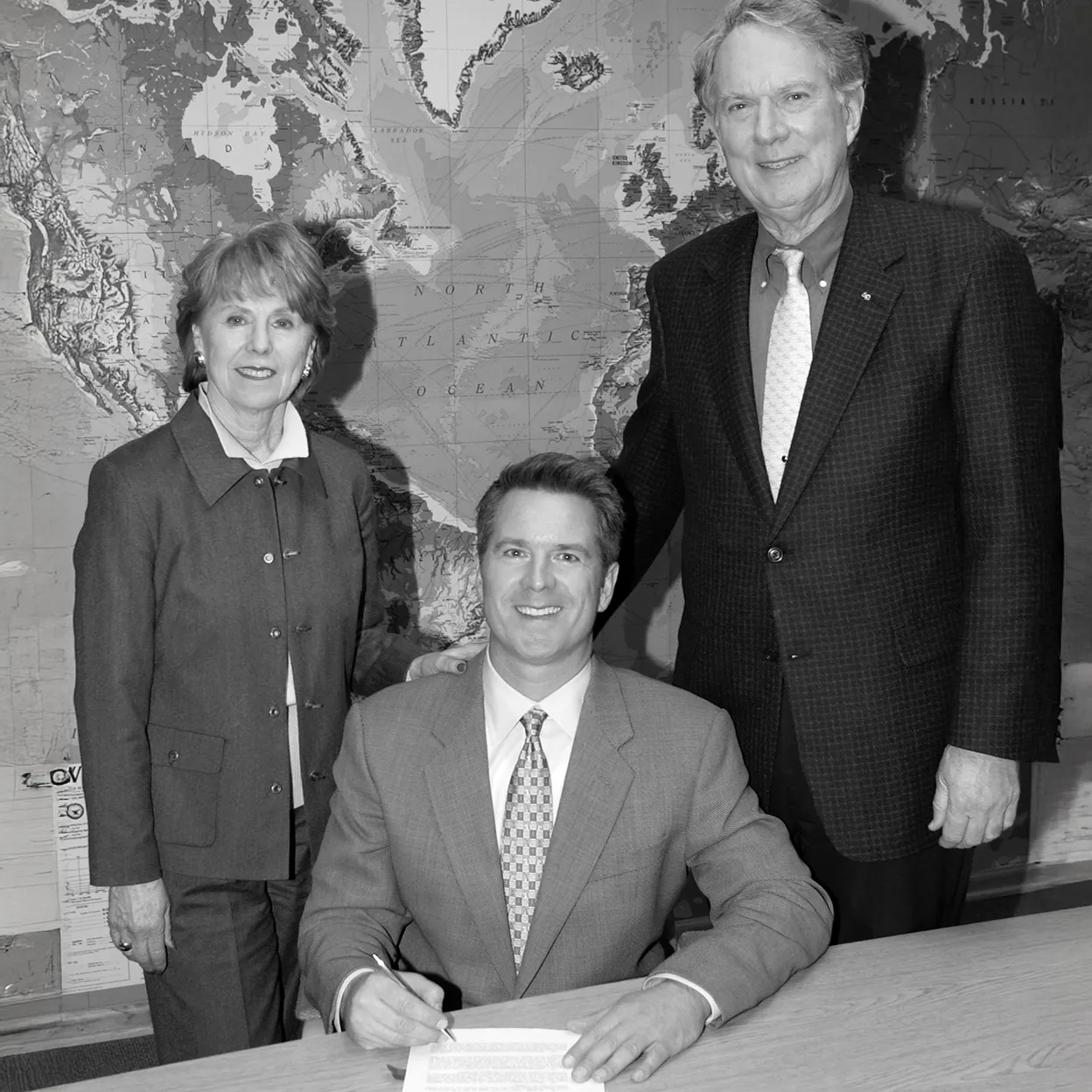 Three business professionals, one seated signing a document and two standing behind, with a large map of the North Atlantic Ocean in the background