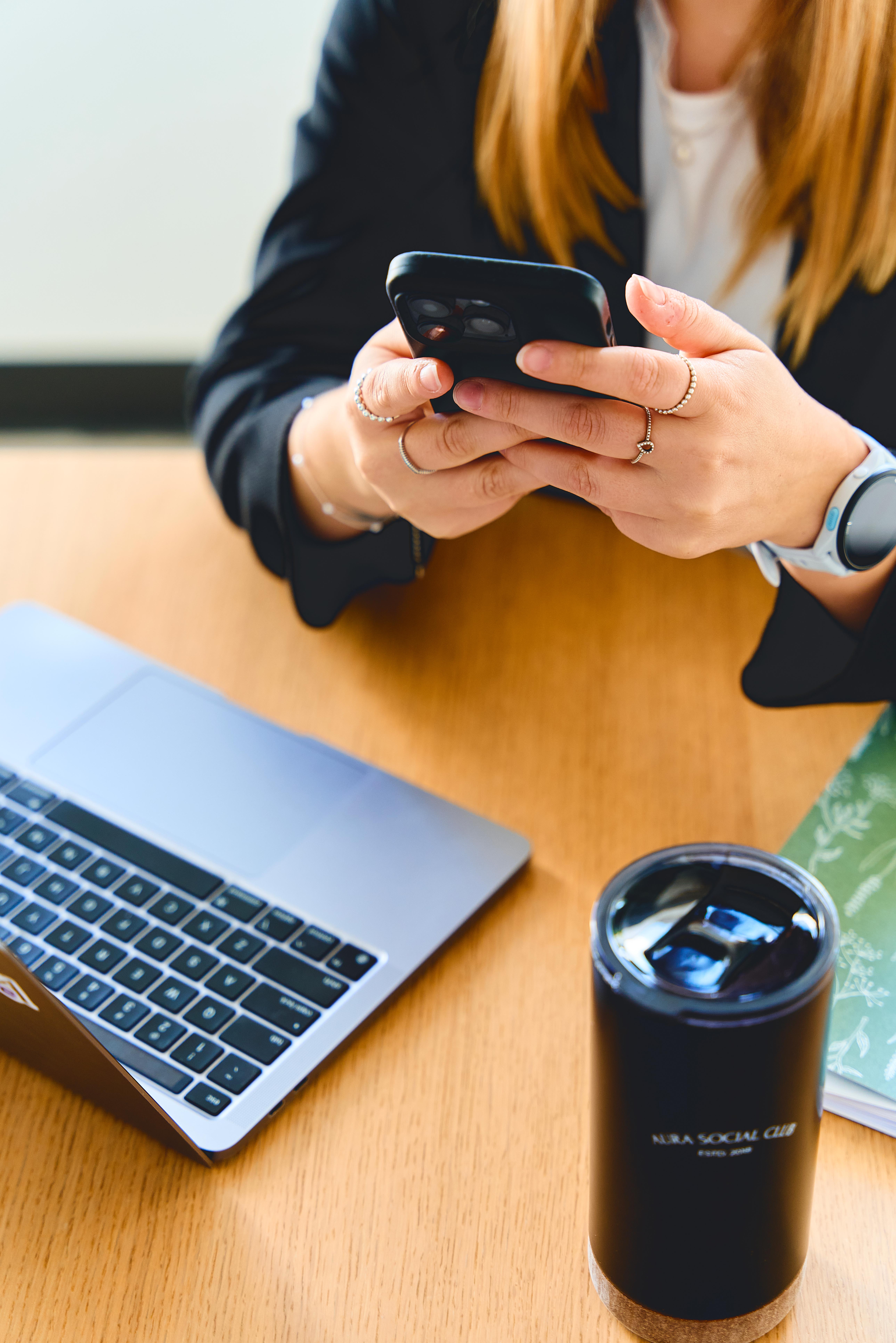 Person with rings on fingers using a smartphone at a desk with a laptop, notebook, and black travel mug.