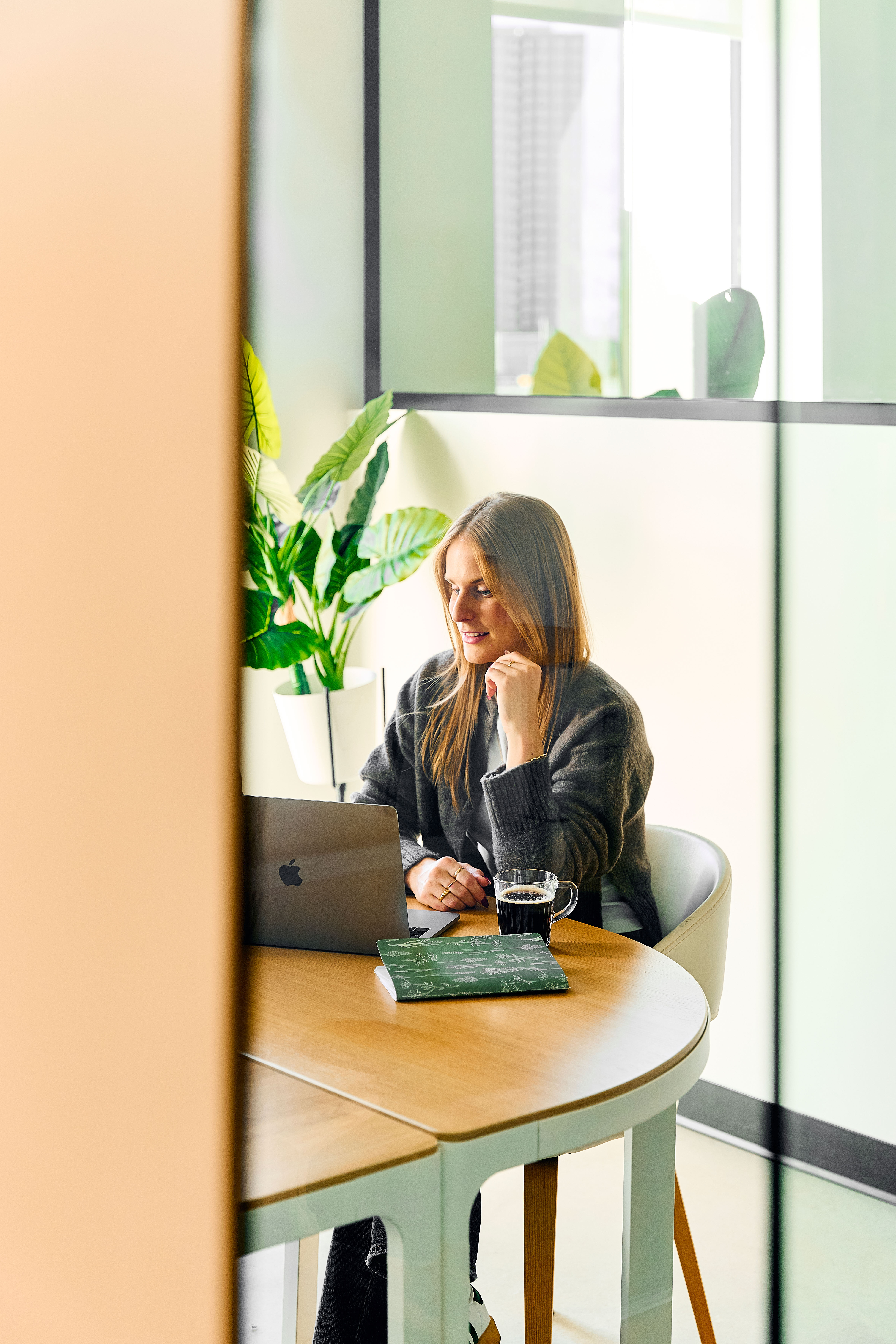 A woman with long blonde hair sitting at a round wooden table using a laptop, with a glass cup of coffee and a green notebook beside her, in a bright modern office with plants.
