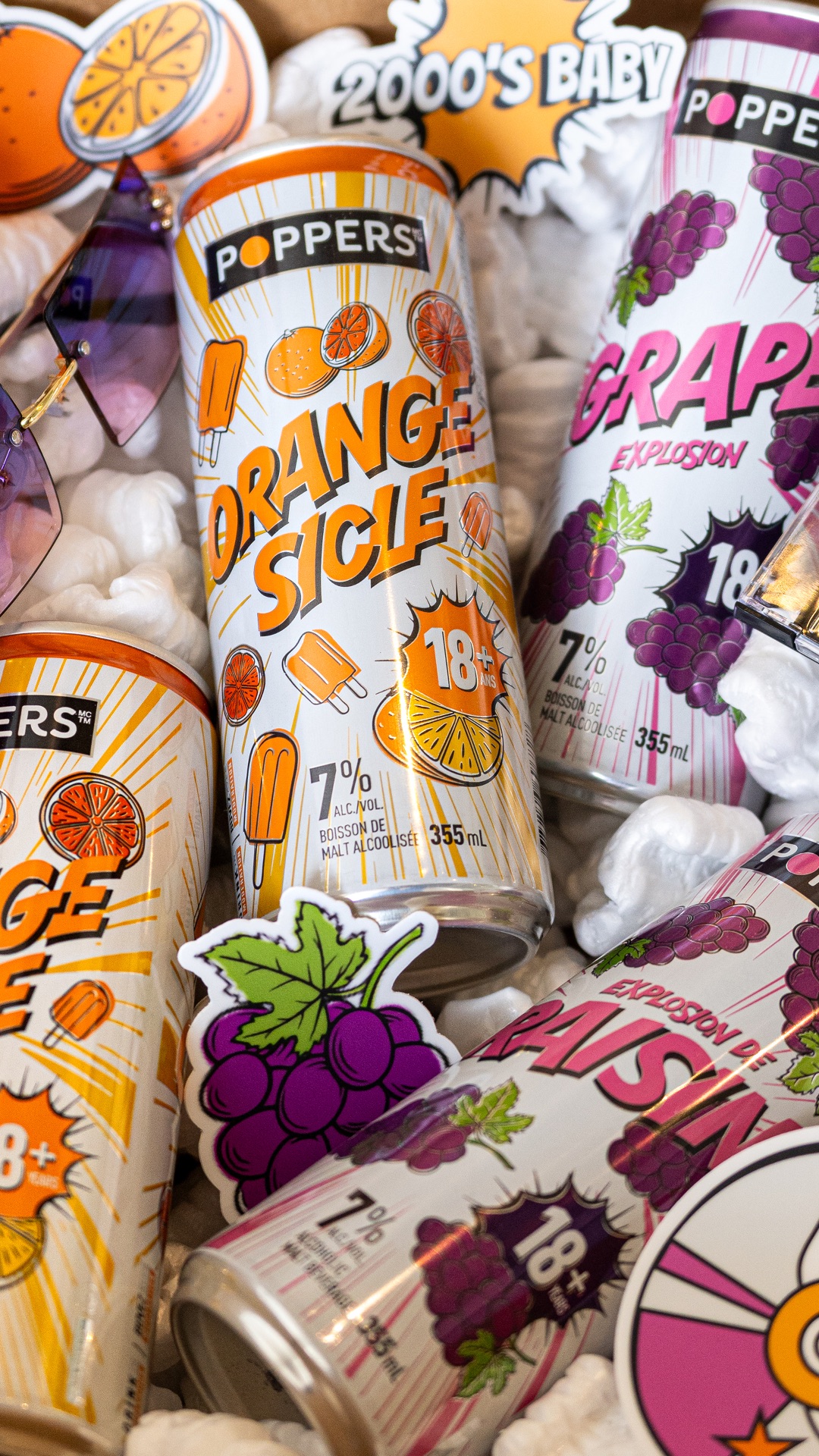 Cans of Poppers alcoholic malt beverages in orange and grape flavors surrounded by popcorn and purple sunglasses.