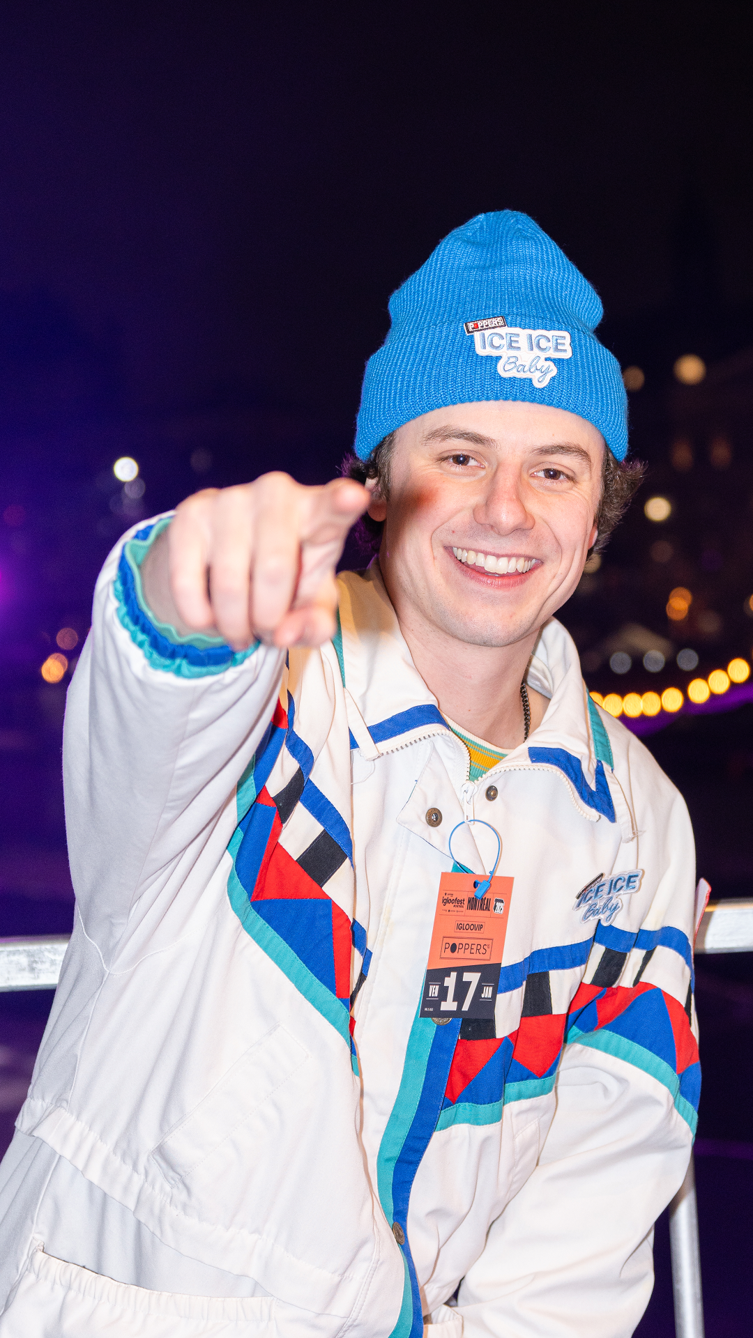 Smiling man wearing a blue beanie and colorful jacket pointing at the camera at night.