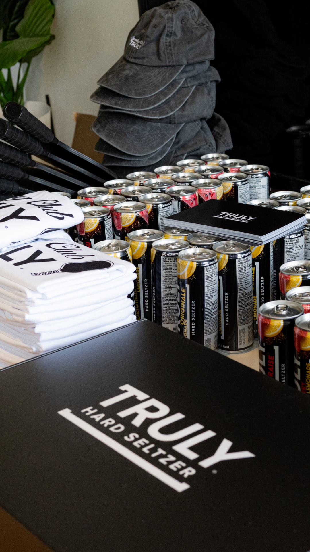 Stacked Truly Hard Seltzer cans surrounded by branded black hats, white t-shirts, and marketing materials on a table.