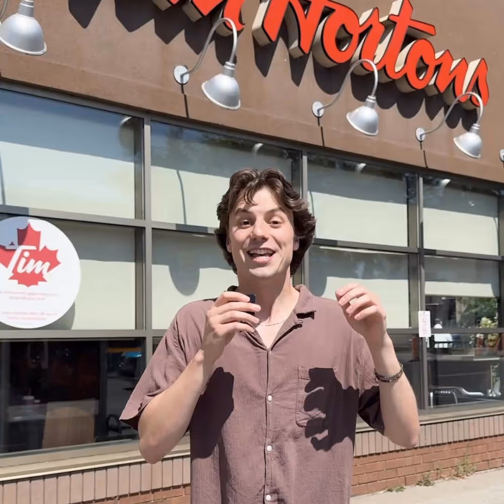 Young man smiling and speaking in front of a Tim Hortons store on a sunny day.