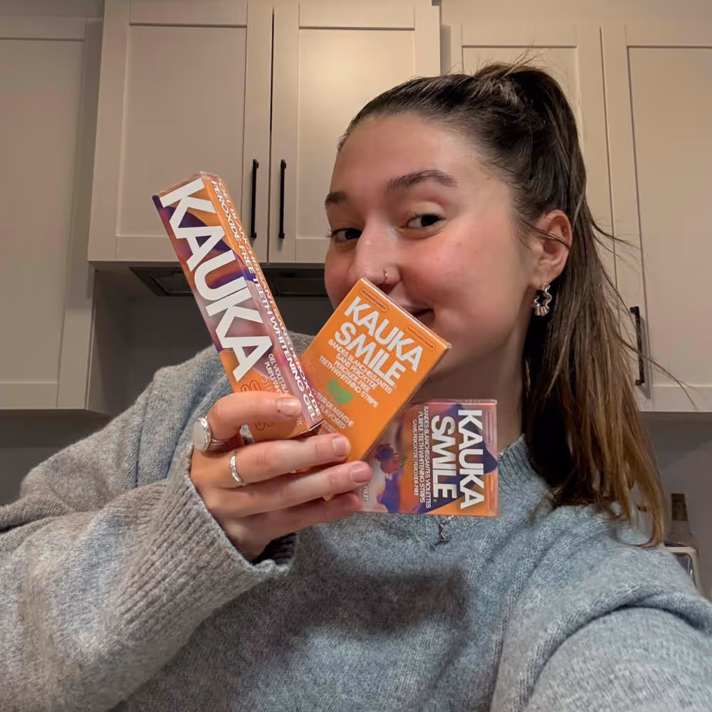 Young woman with long hair and gray sweater holding up Kauka Smile teeth whitening products in a kitchen.