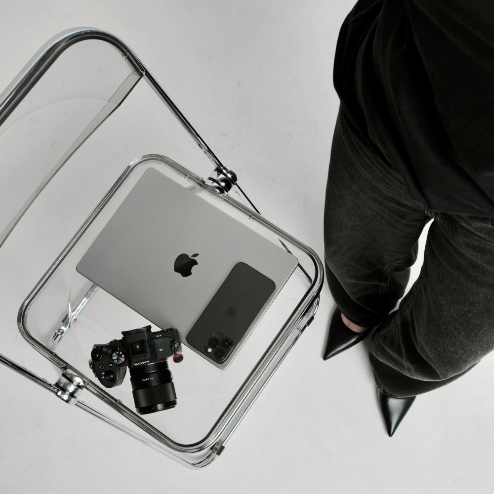 Top view of a clear chair holding a MacBook, an iPhone, and a Sony camera next to a person wearing black pointed shoes and dark pants.