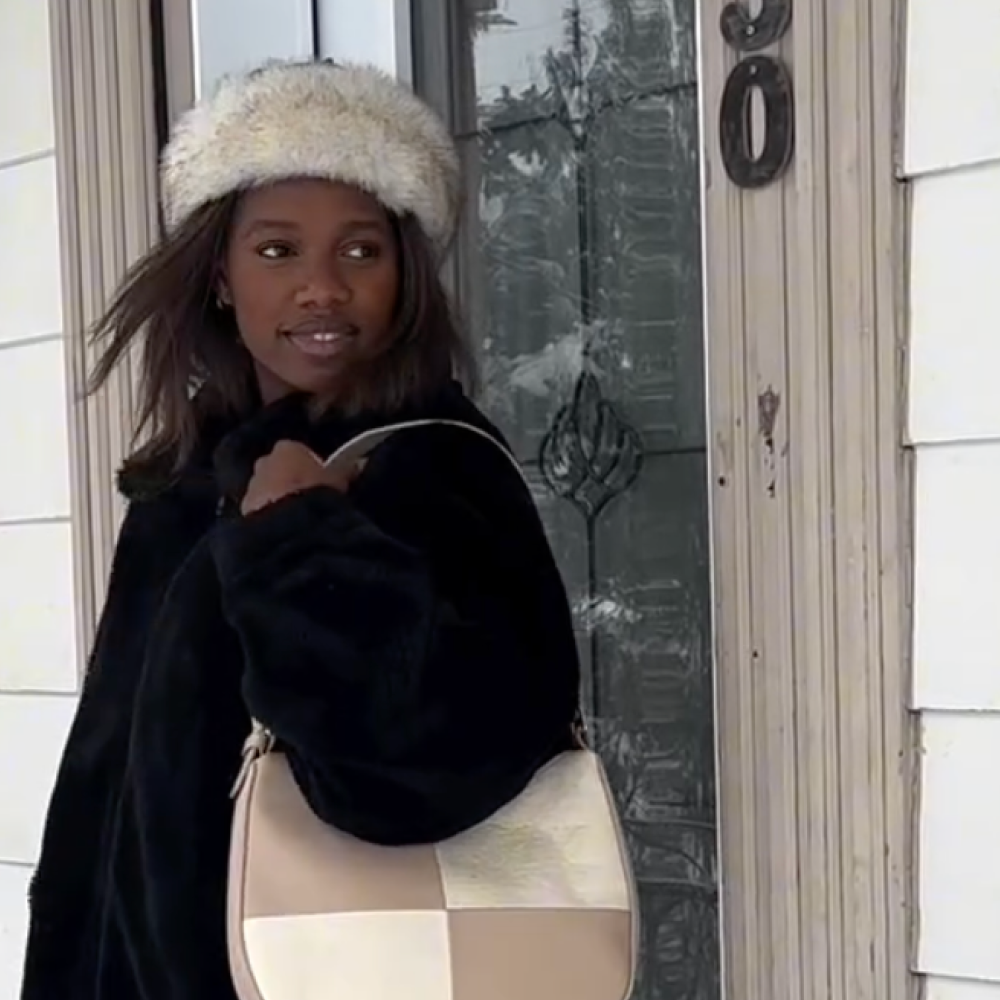 Woman wearing a fluffy white hat and black coat, carrying a beige and white patchwork handbag, standing near a decorative glass door.