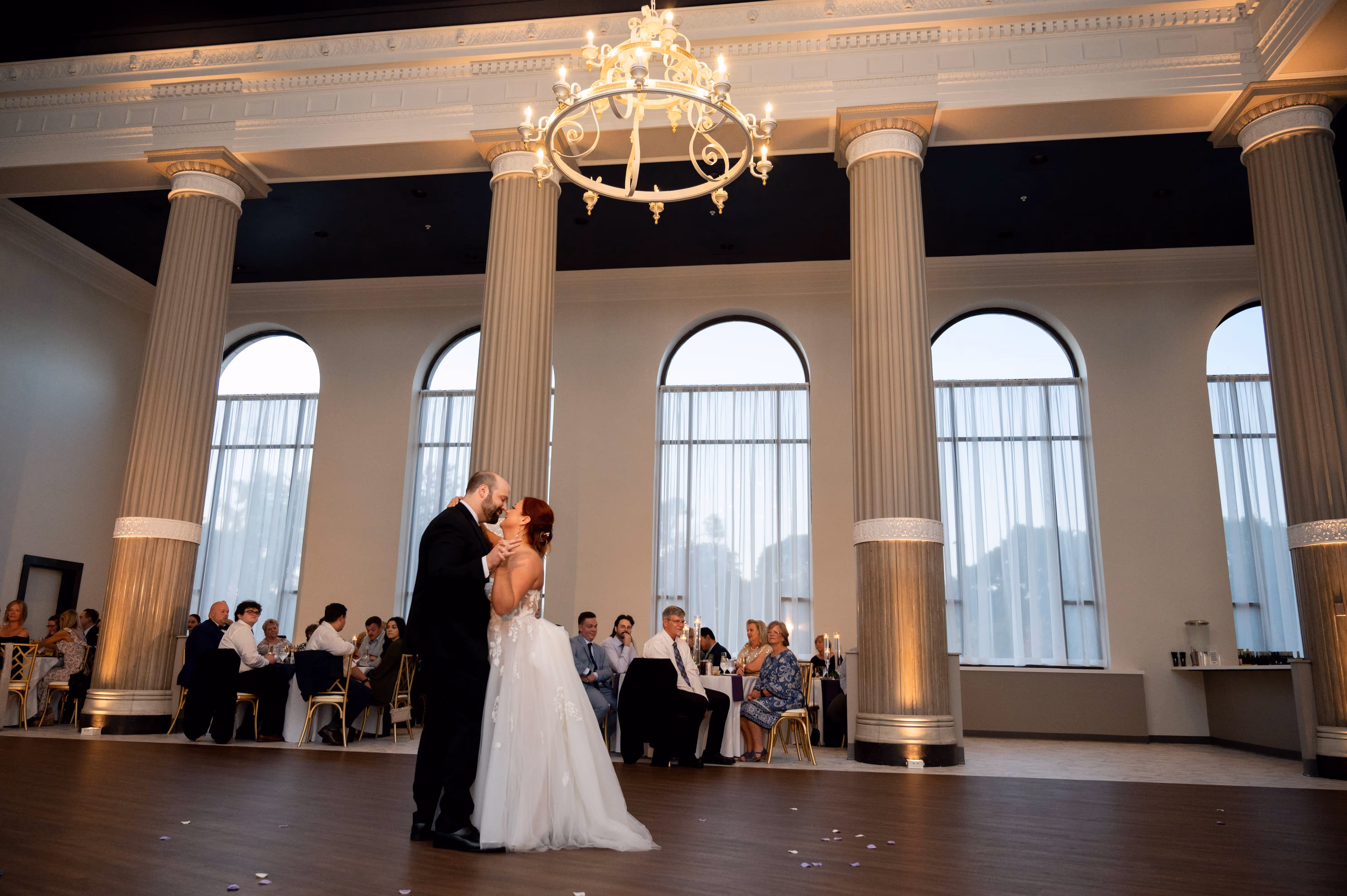Bride and groom sharing a first dance inside The Bank at East End, a room with large arched windows and tall columns, and guests seated in the background.