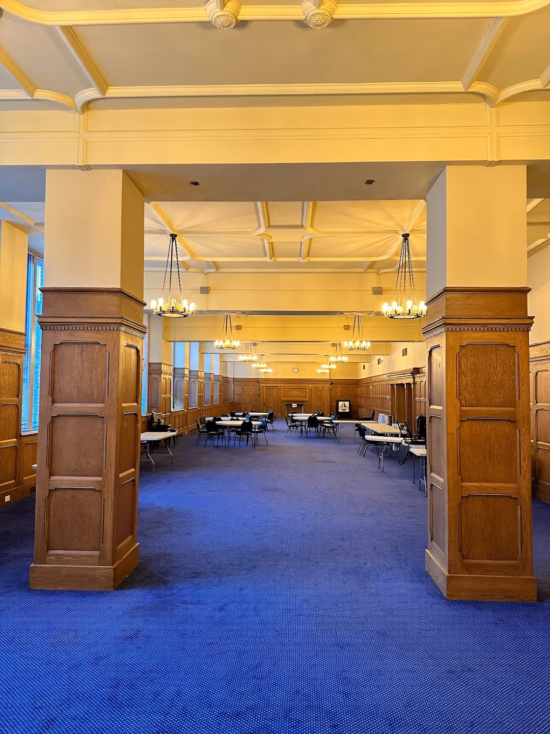 The Community Room at East End with blue carpet, wooden paneling on walls and columns, round chandeliers, and tables with chairs arranged around the perimeter.