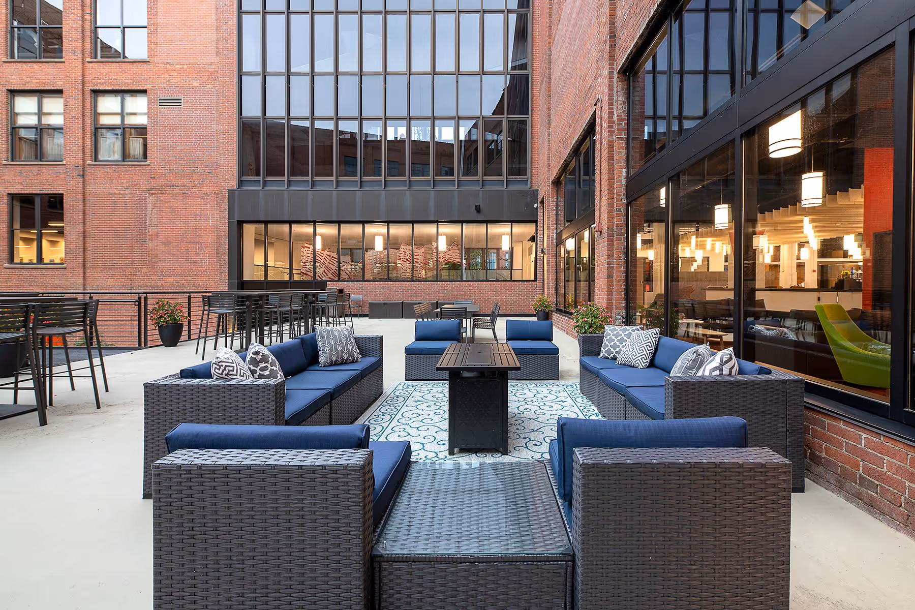 Outdoor patio seating area with blue cushioned wicker sofas and chairs around a black fire pit table on a patterned rug, surrounded by brick buildings with large windows.