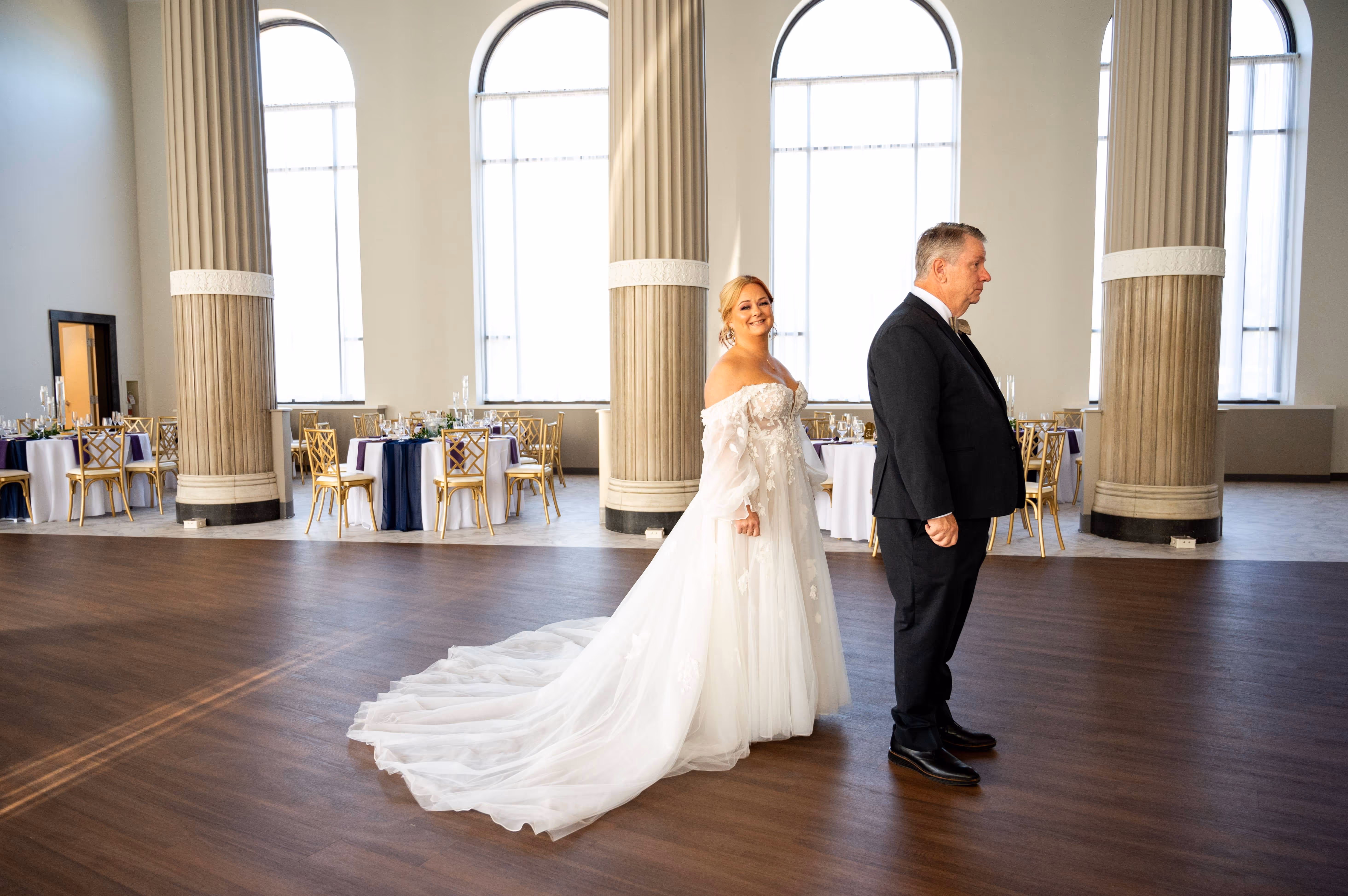Bride in a white wedding gown smiling behind a man in a black suit standing inside The Bank at East End, a room with tall columns and banquet tables.