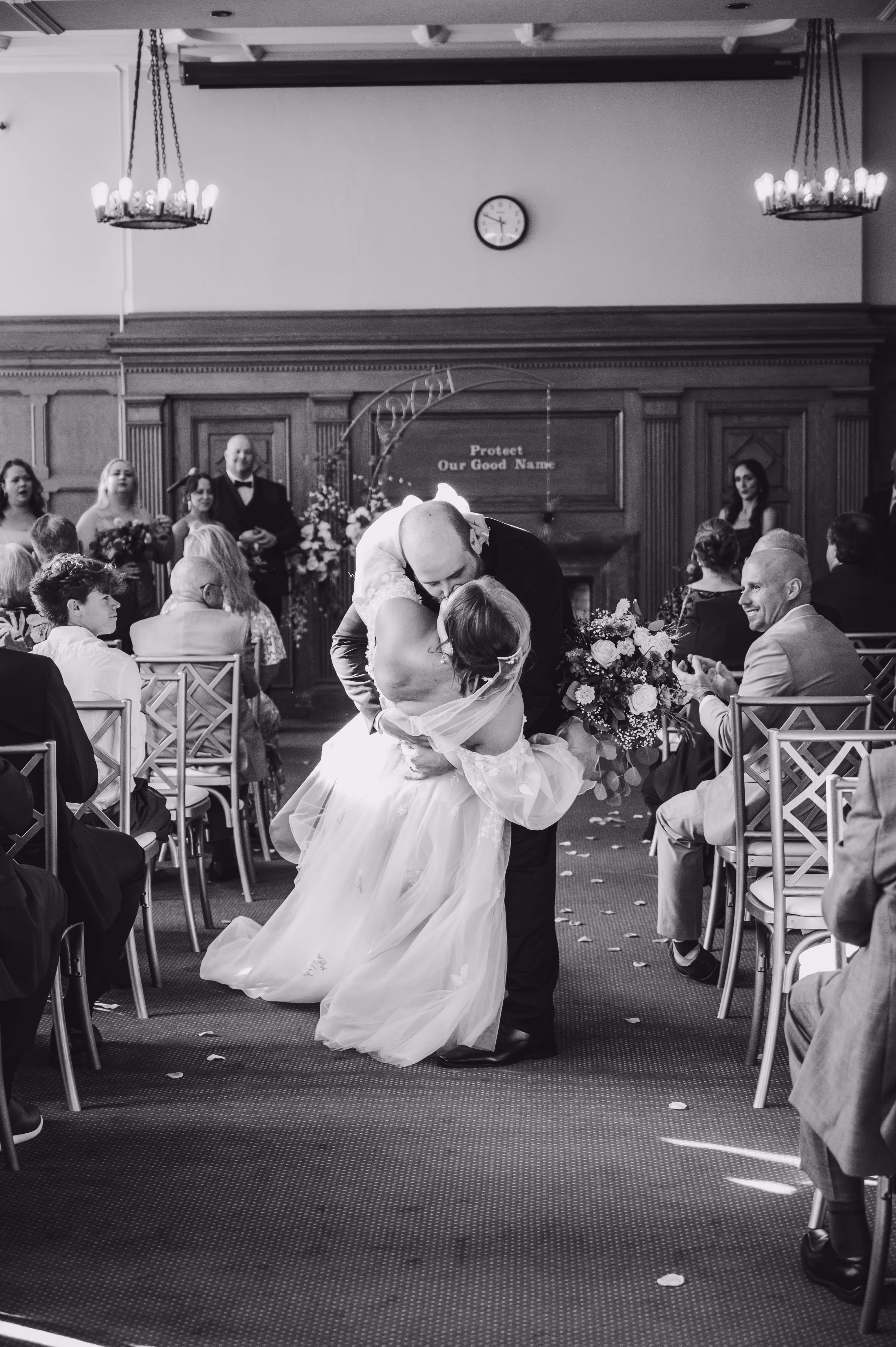Bride and groom sharing a romantic kiss during a wedding ceremony inside The Community Room at East End, a room with seated guests and wooden paneling.