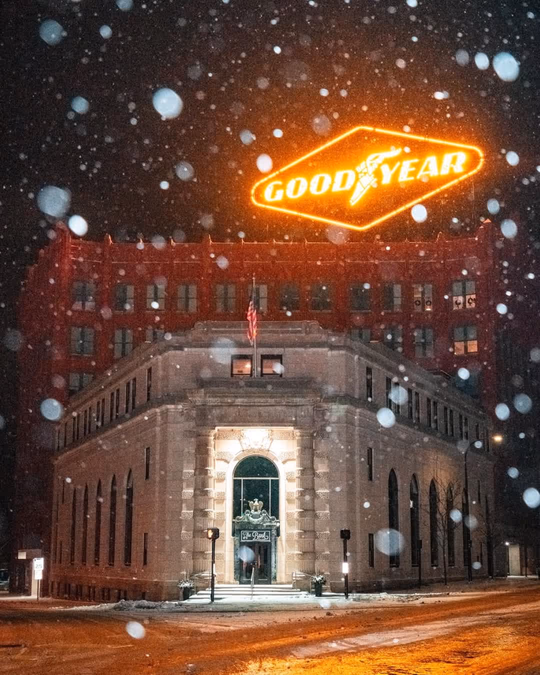 Night view of the historic Bank at East End building with snow falling and the illuminated Goodyear sign glowing above.